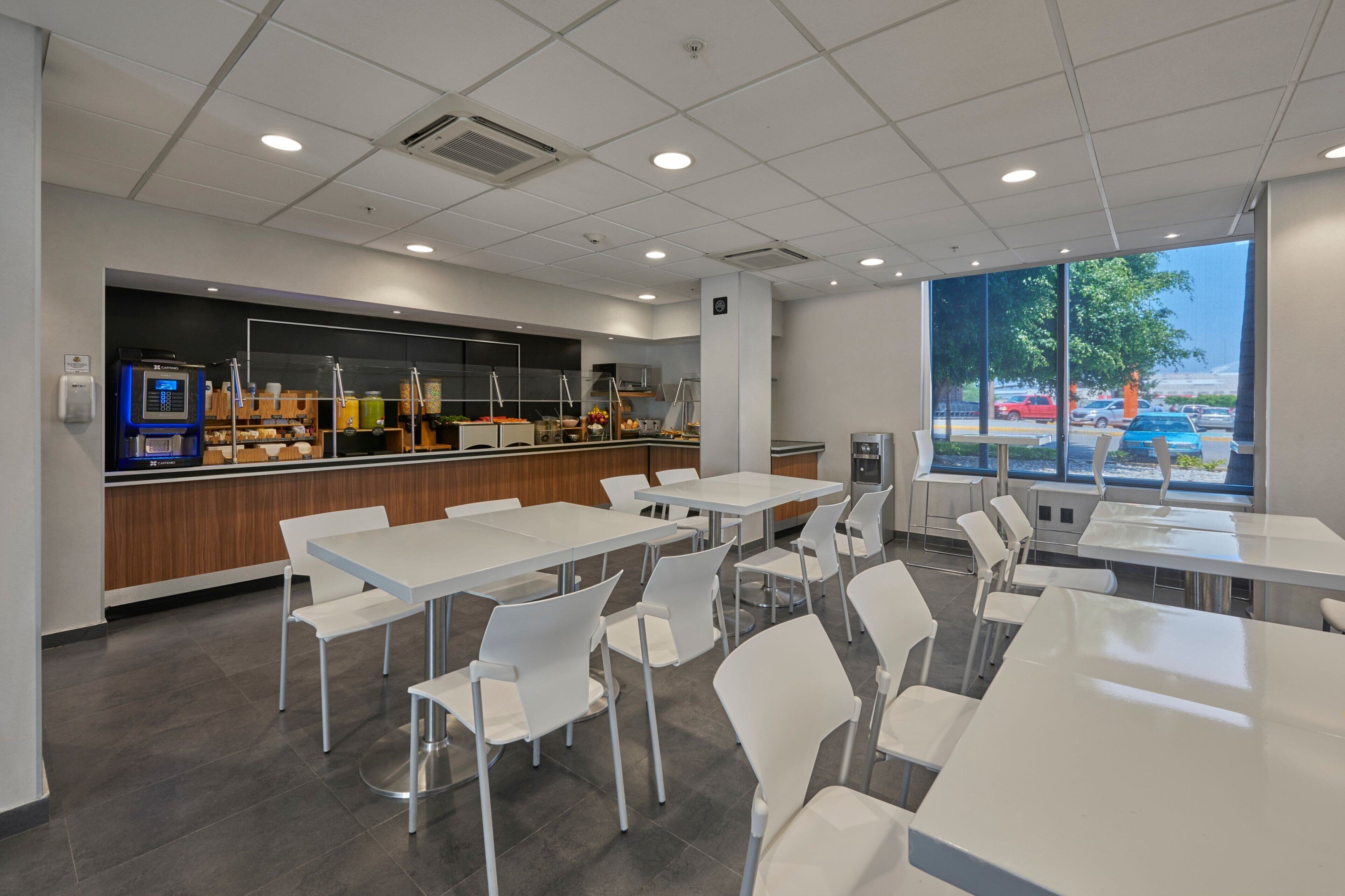 Una cafetería moderna con mesas y sillas blancas, una estación de autoservicio para bebidas y ventanas amplias con vistas a coches aparcados y árboles.