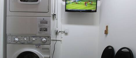 A laundry room with a washing machine, a dryer, a television mounted on the wall, and a chair.