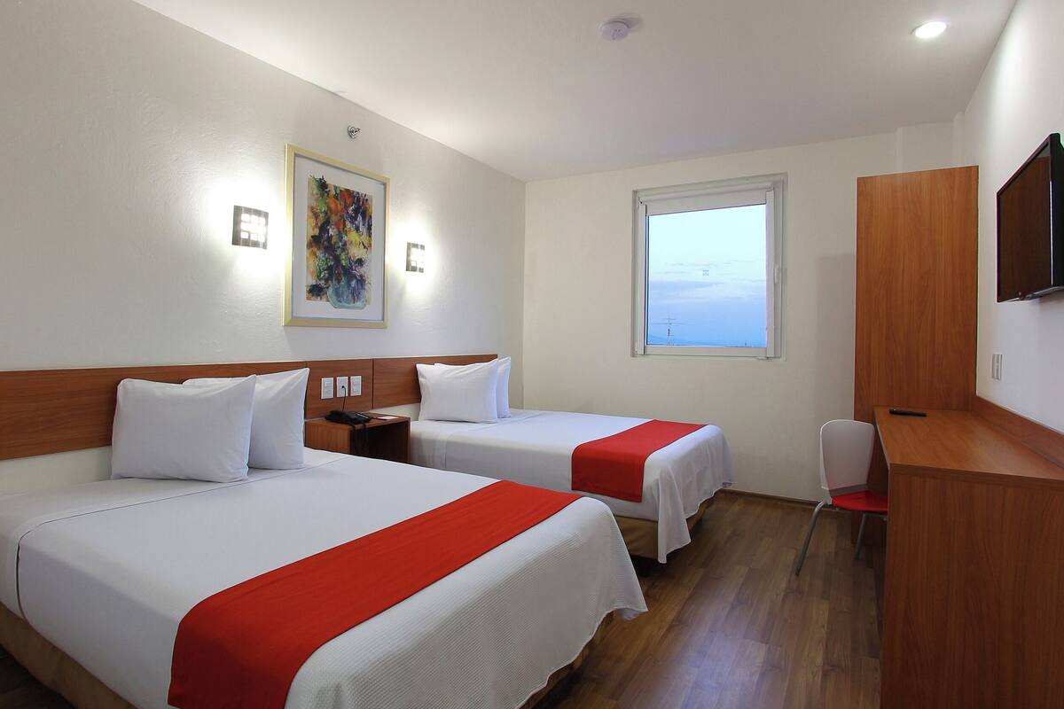 Ein Hotelzimmer mit zwei Betten, einem Schreibtisch, einem Stuhl, einem Fenster und einem Gemälde an der Wand.