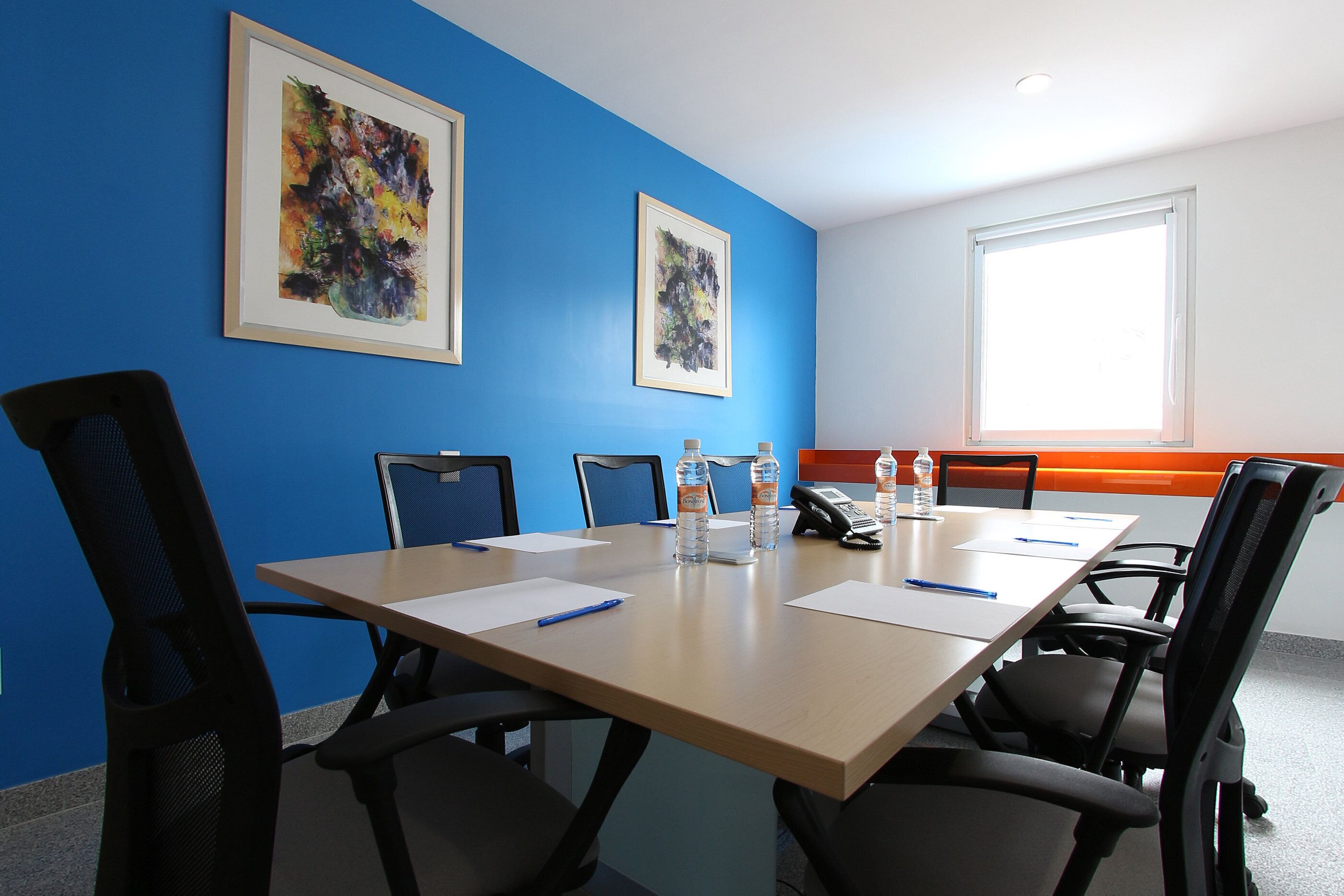 Een vergaderruimte met een lange tafel, stoelen, waterflesjes en lijstjes met kunstwerken aan de muren.