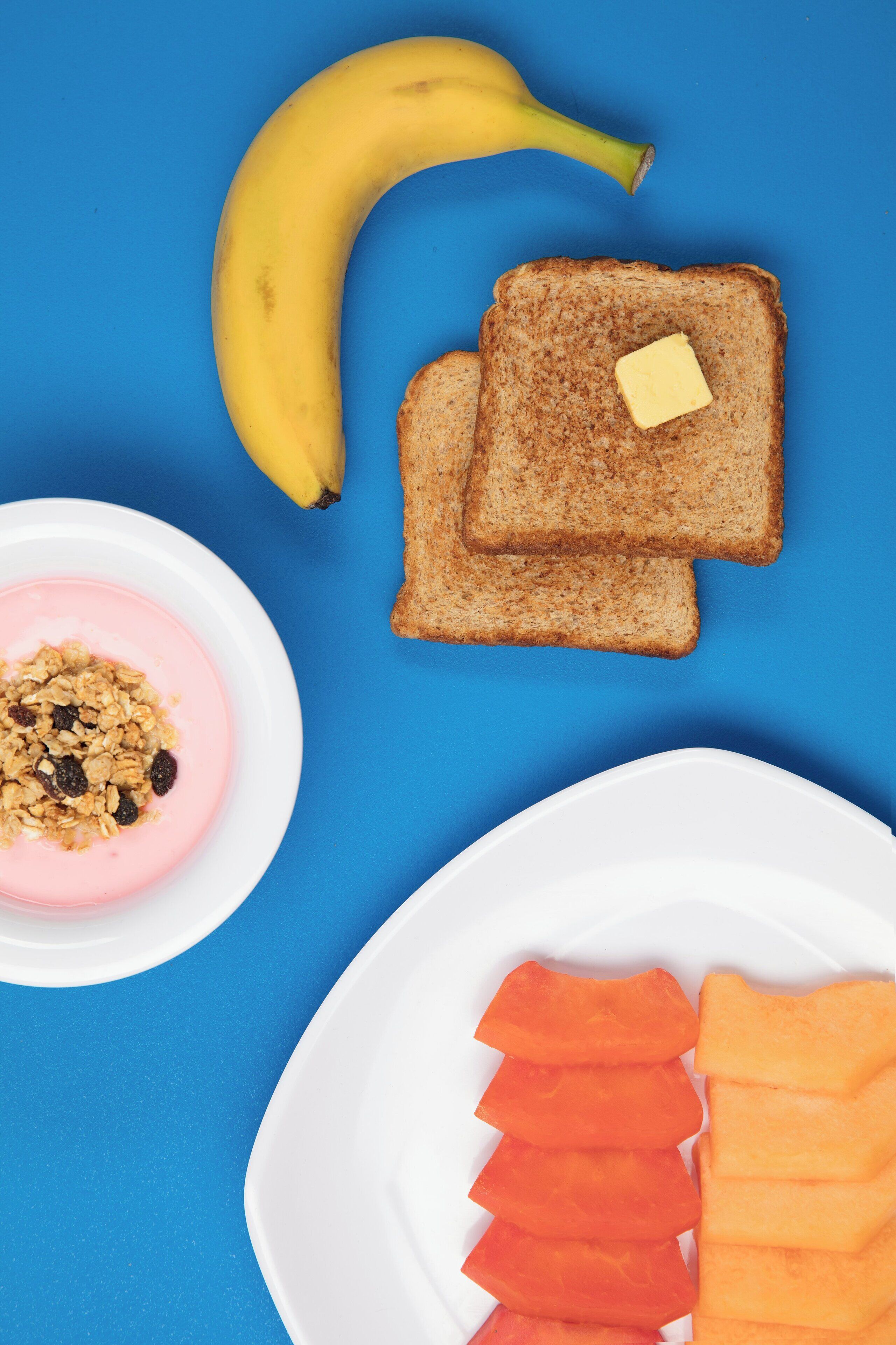 A breakfast setting with toast, a banana, yogurt with granola, and sliced fruit on a blue table.