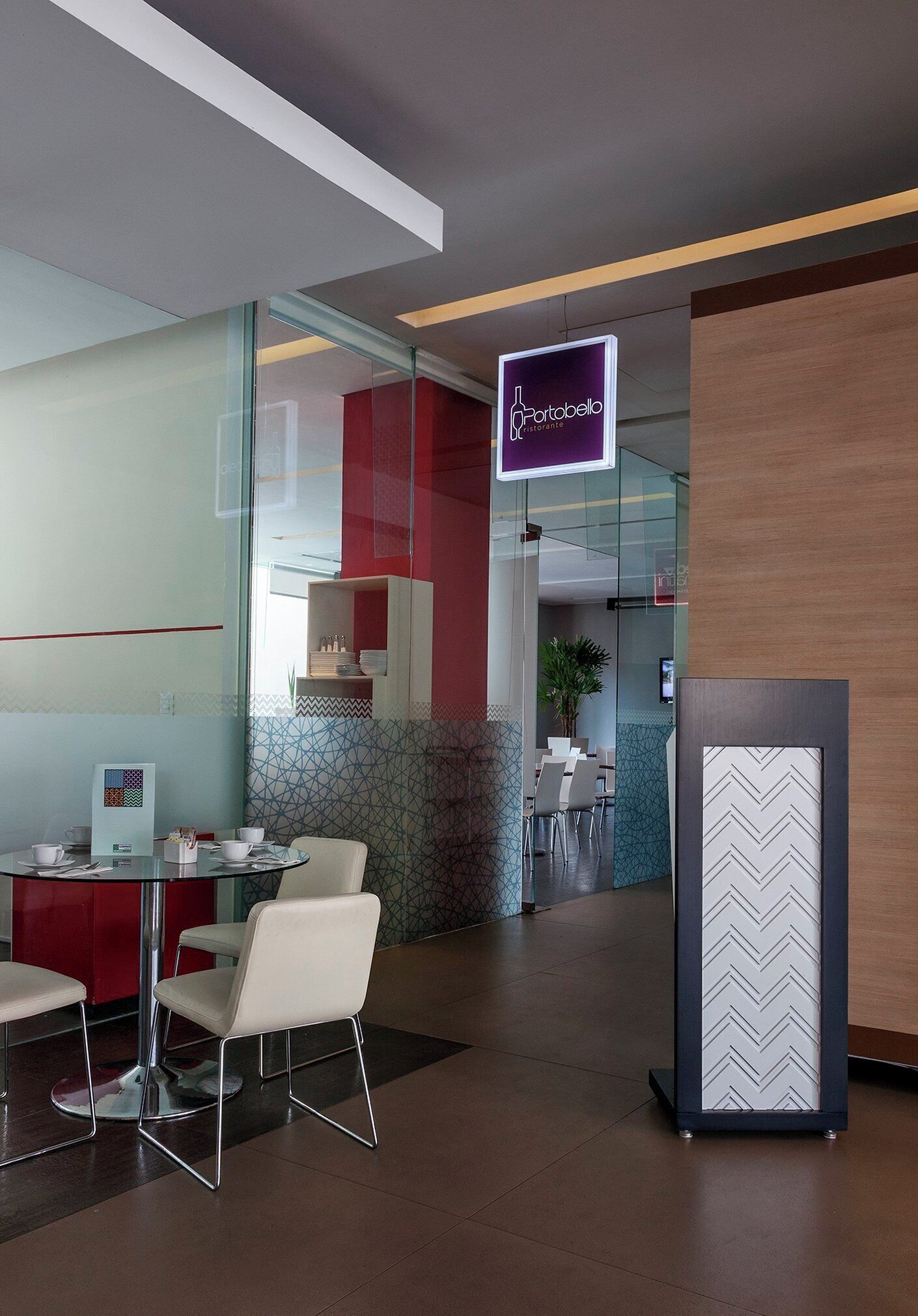 A modern hotel lobby with a reception desk, seating area, and a glass partition leading to a dining area.