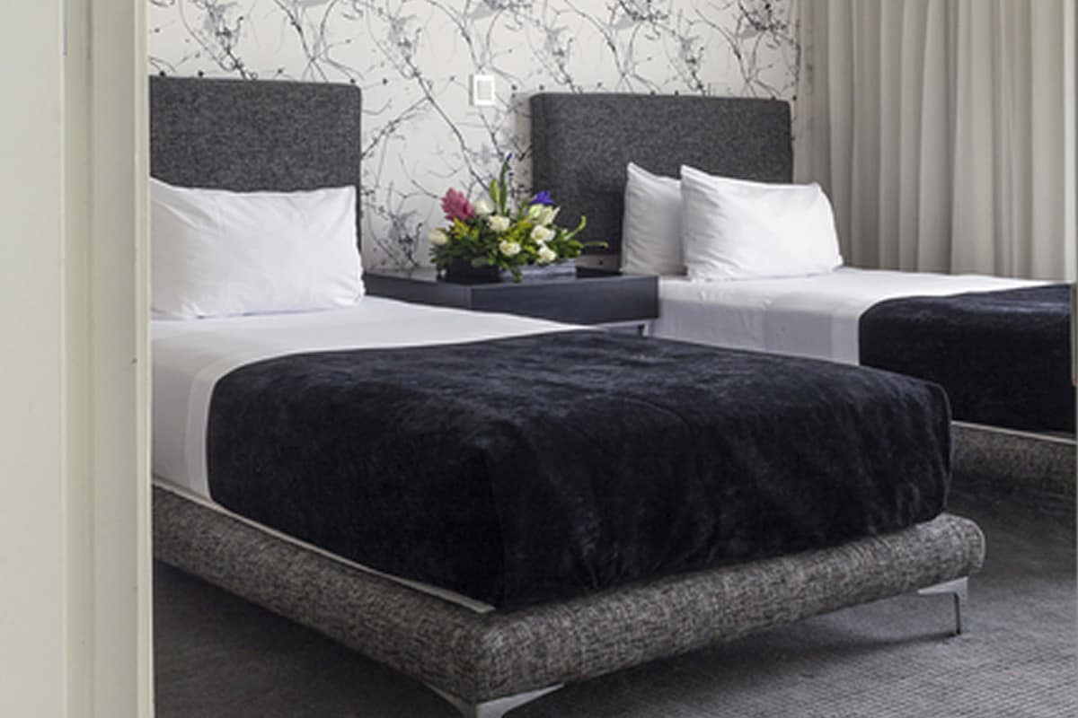 A hotel room with two beds, each with a grey headboard and white bedding. There is a small table with a vase of flowers between the beds.