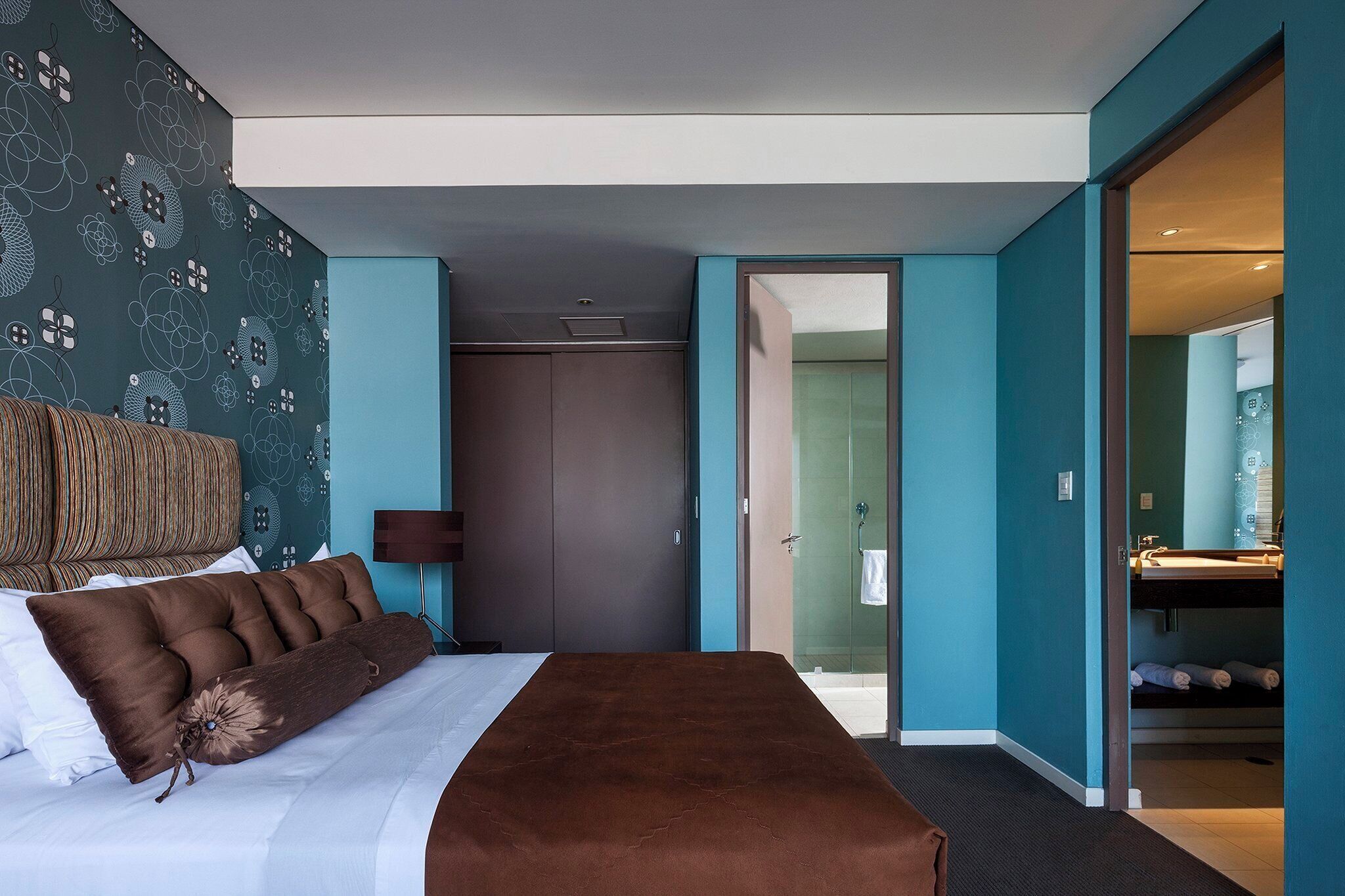 A modern hotel room with a bed, a large window, a bathroom with a sink, and a door leading to another room.