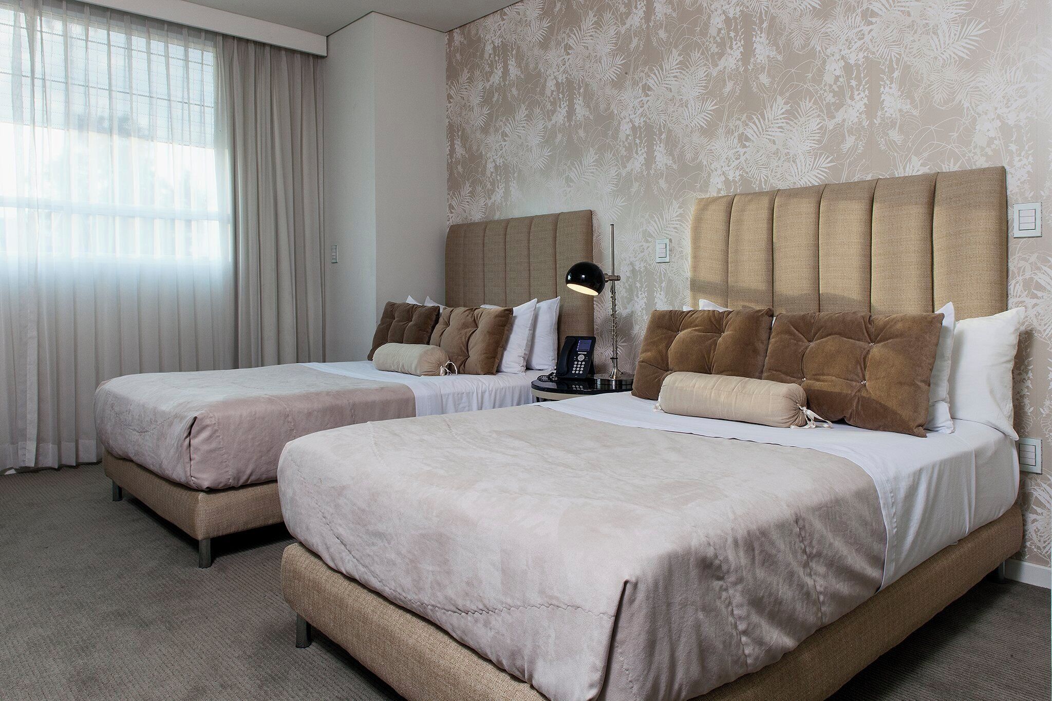 A hotel room with two beds, a headboard, a bedside table with a lamp, and a window with curtains.