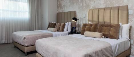 A hotel room with two beds, a headboard, a bedside table with a lamp, and a window with curtains.