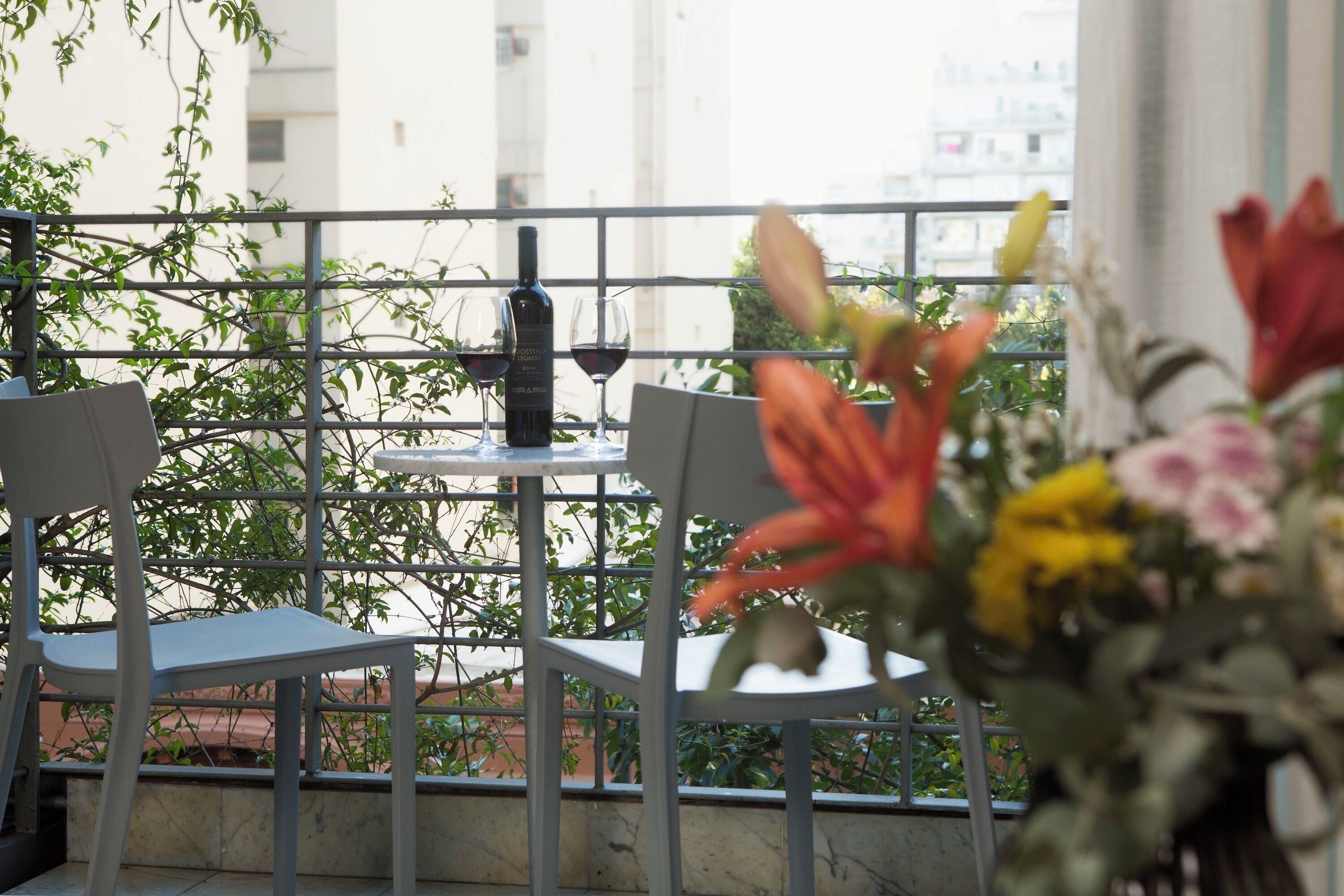 A balcony with two chairs, a table with a bottle and glasses, and a vase of flowers.