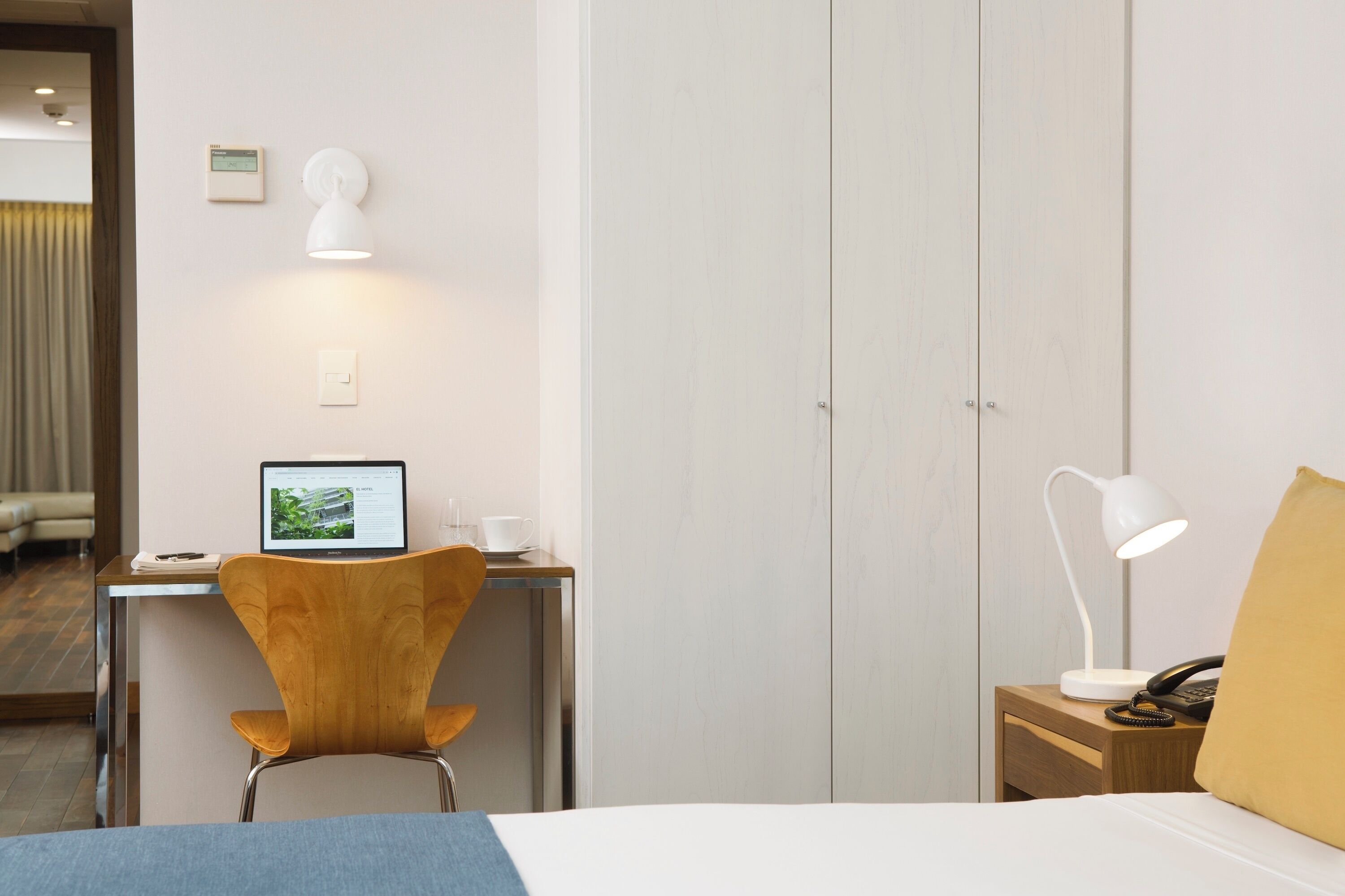 A hotel room with a bed, a desk with a laptop, a chair, a bedside table with a lamp, and a wardrobe.