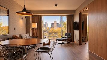 A modern hotel room with a dining area, a sofa, a TV, and a view of the cityscape.