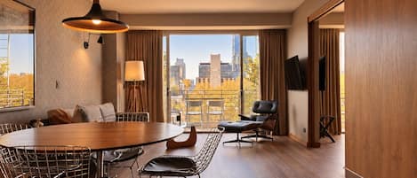 Habitación de hotel moderna con zona de comedor, sofá, televisor y vista panorámica de la ciudad.