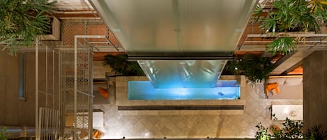 A modern indoor pool area with a glass ceiling, surrounded by plants and a stone wall.