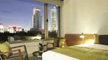 A hotel room with a bed, a chair, a small table with a plant, and a view of the cityscape through large windows.