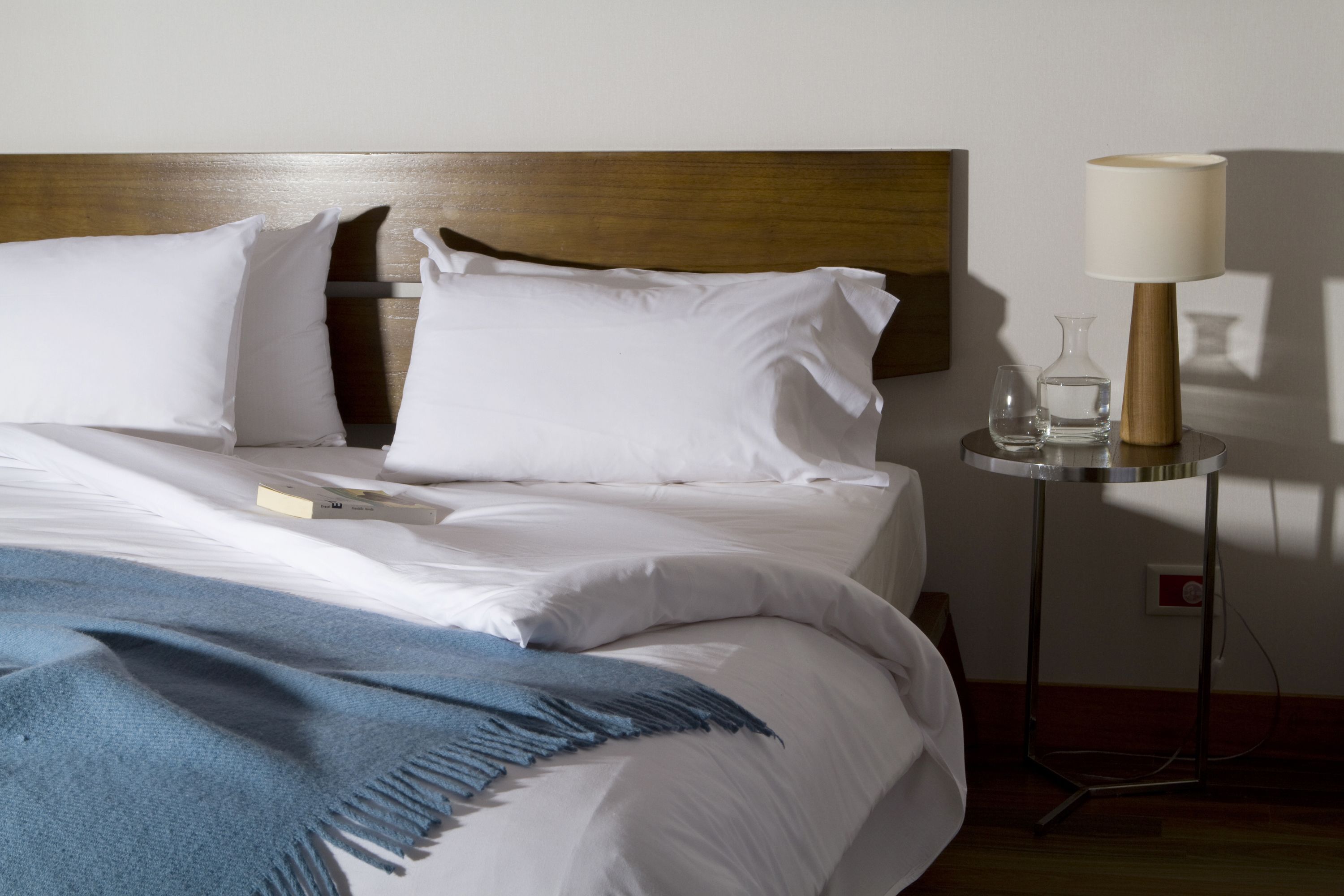 A neatly made bed with white linens and a blue throw blanket, a bedside table with a lamp and a glass vase, and a wooden headboard.