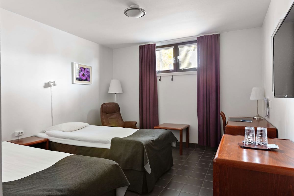 Een hotelkamer met twee bedden, een bureau, een stoel, een lamp en een raam met gordijnen.