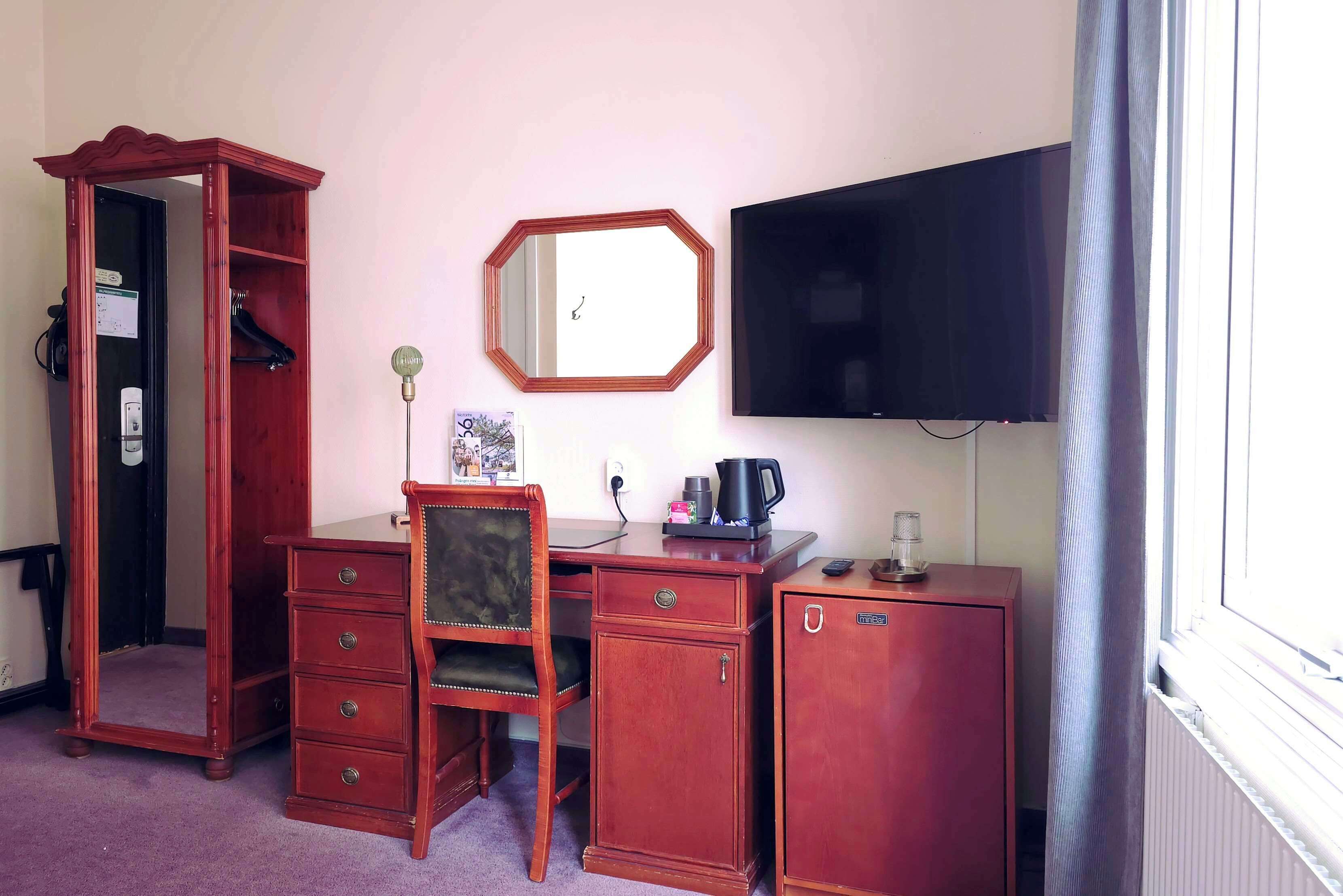 Una habitación con un escritorio de madera, una silla, un armario, un espejo, un televisor y un radiador.