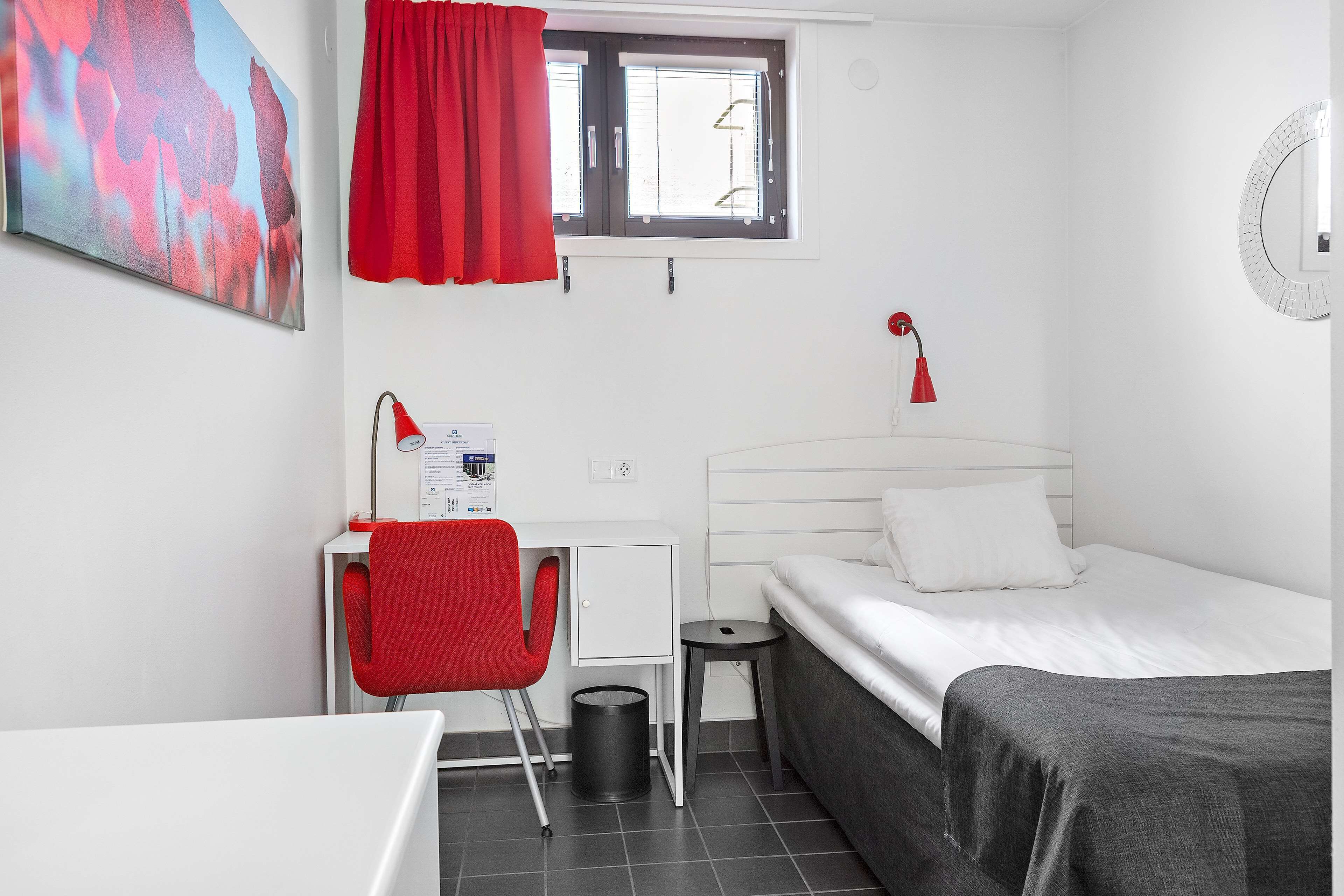 Una habitación pequeña y limpia con una cama, un escritorio con una silla, una ventana con cortinas y una lámpara fijada a la pared.