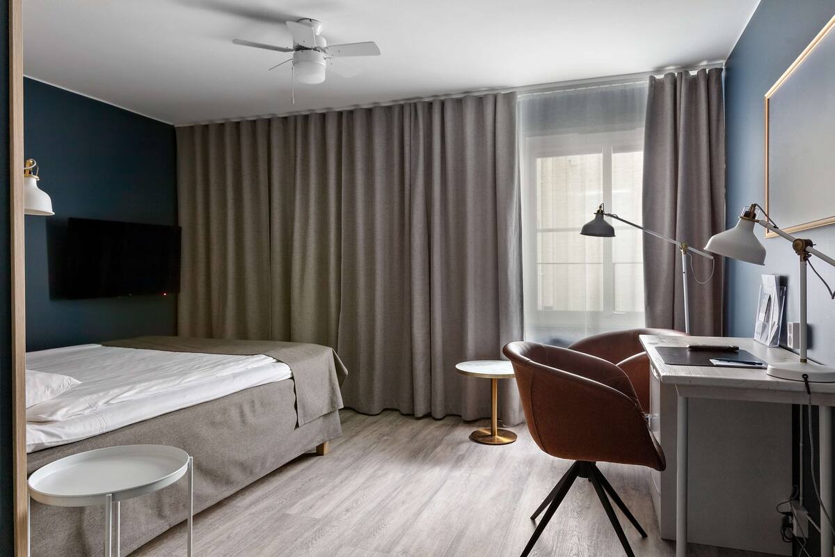 Een moderne hotelkamer met een bed, bureau, stoel, plafondventilator en een aan de muur bevestigde televisie.