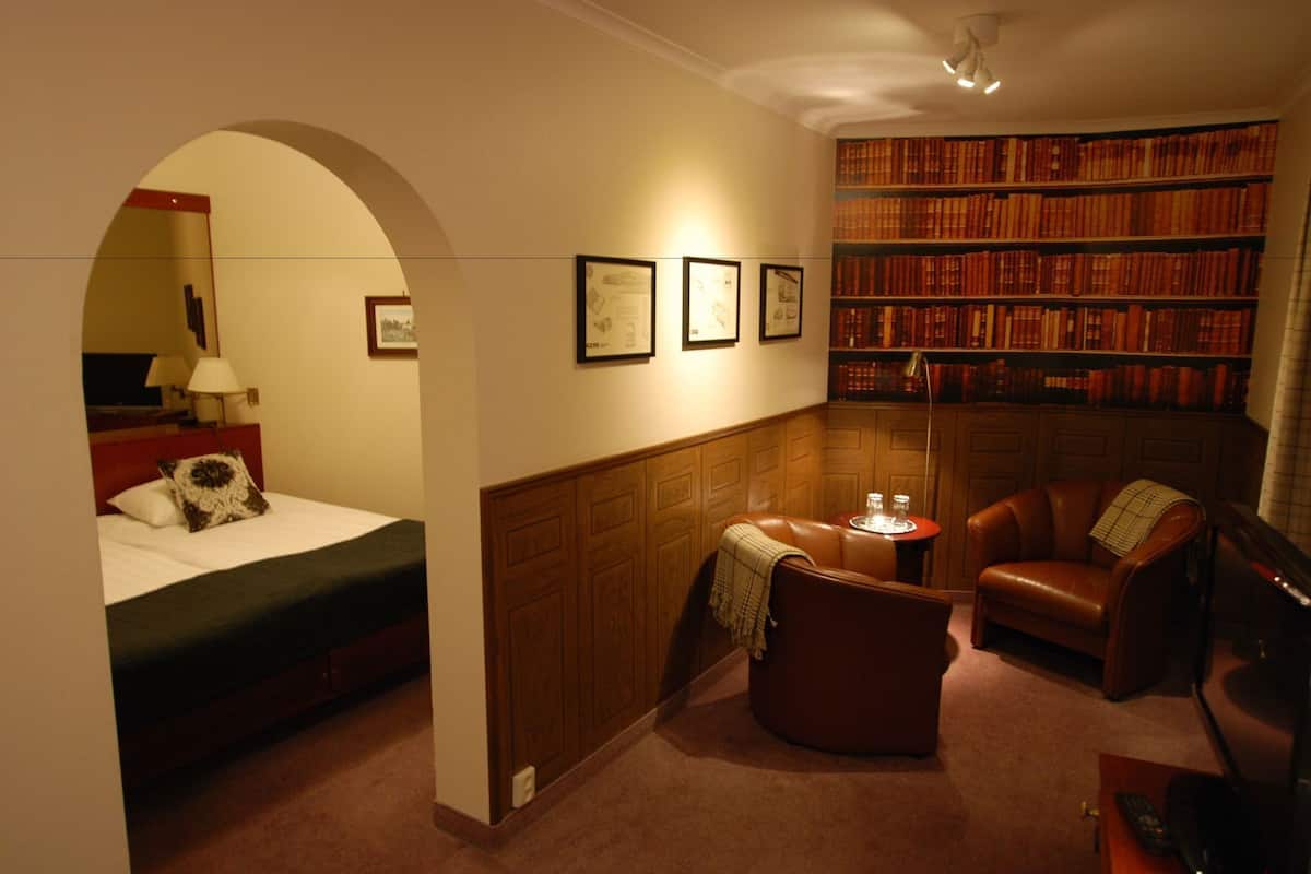 Een hotelkamer met een bed, een klein tafeltje met glazen, een leren fauteuil, een boekenplank en houten wandpanelen.