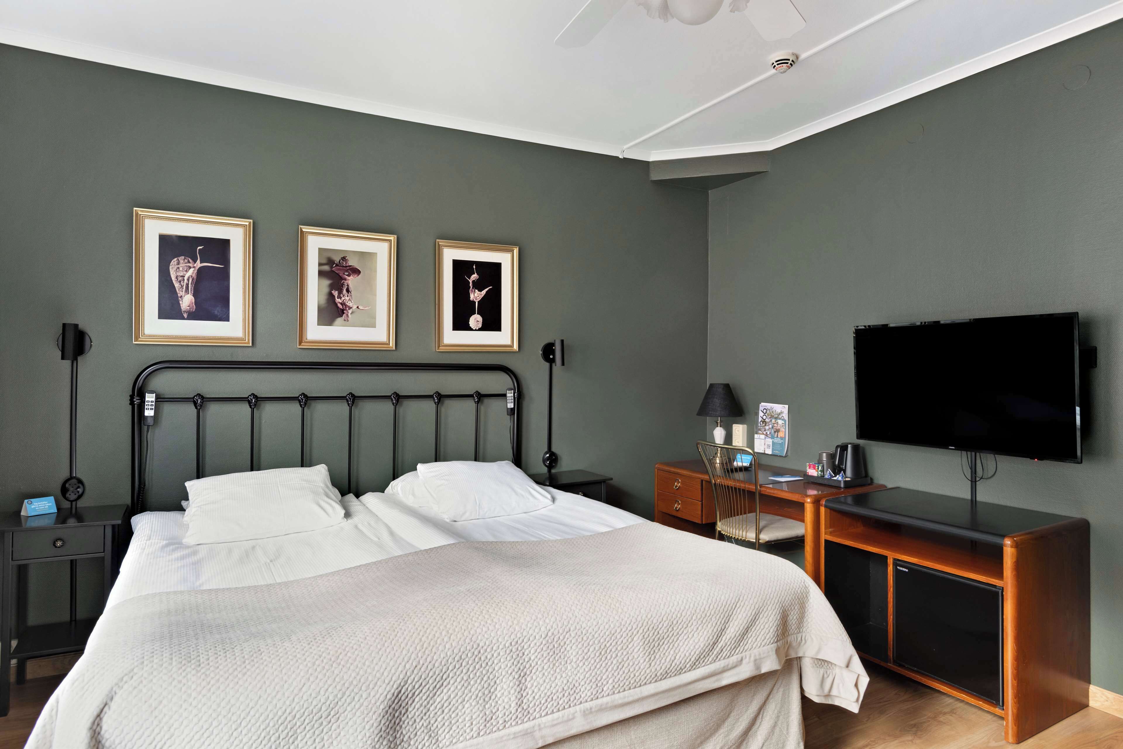 A neatly arranged bedroom with a bed, bedside tables, a desk, a chair, and a flat-screen TV mounted on the wall.