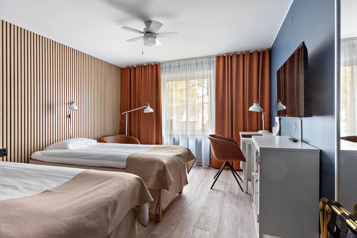 Een hotelkamer met twee bedden, een bureau, een stoel, een plafondventilator en een groot raam met gordijnen.