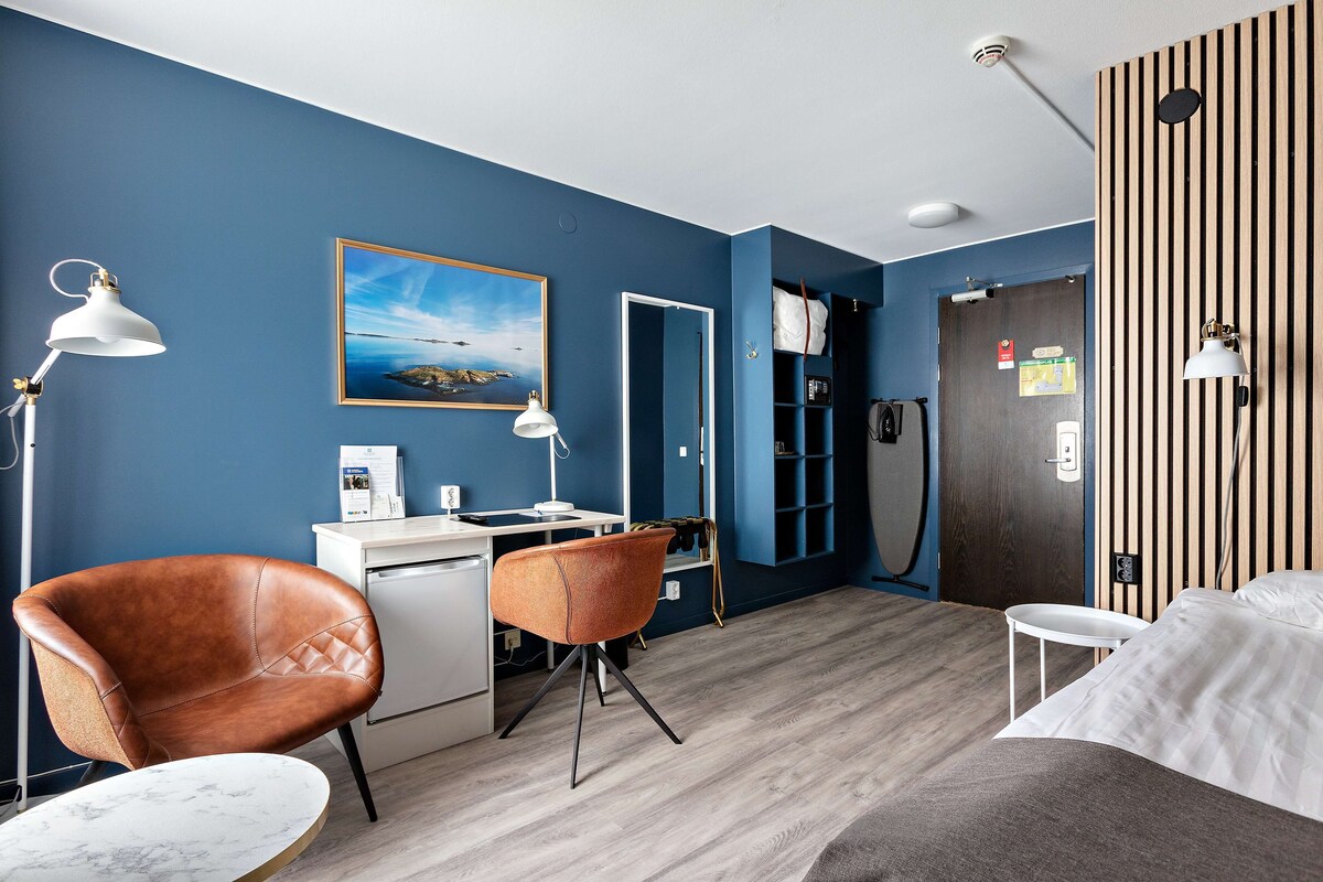 A room with a desk, two chairs, a bed, and a wooden wall with vertical stripes.