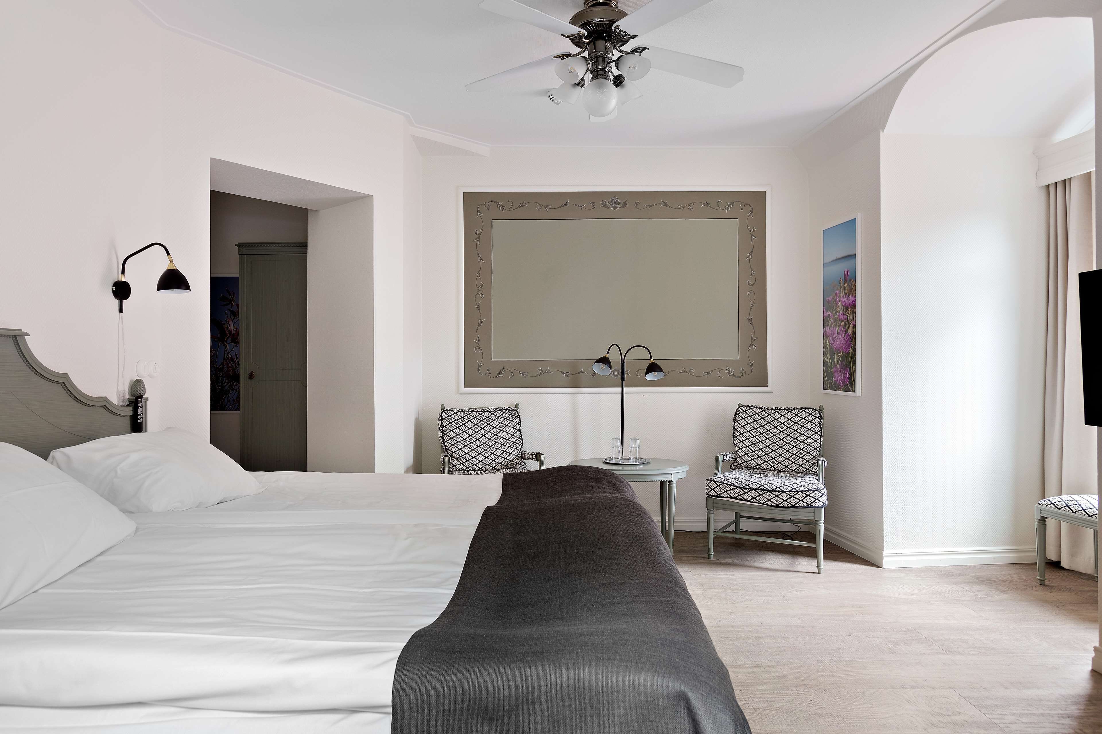 A hotel room with a bed, two chairs, a table, a ceiling fan, and a large mirror.