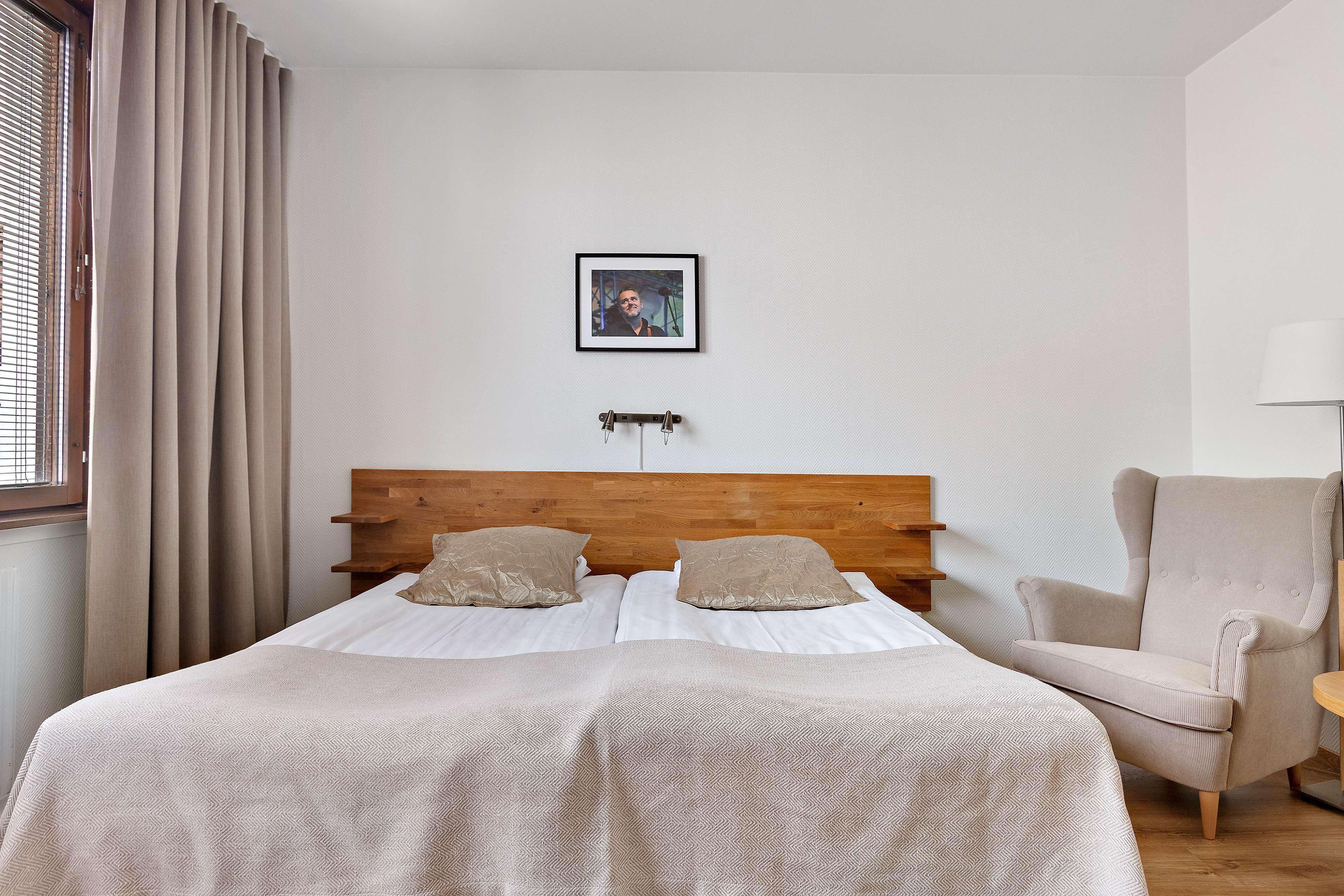 Un dormitorio ordenado con cabecera de madera, dos camas con ropa de cama color beige, una silla y un cuadro en la pared.