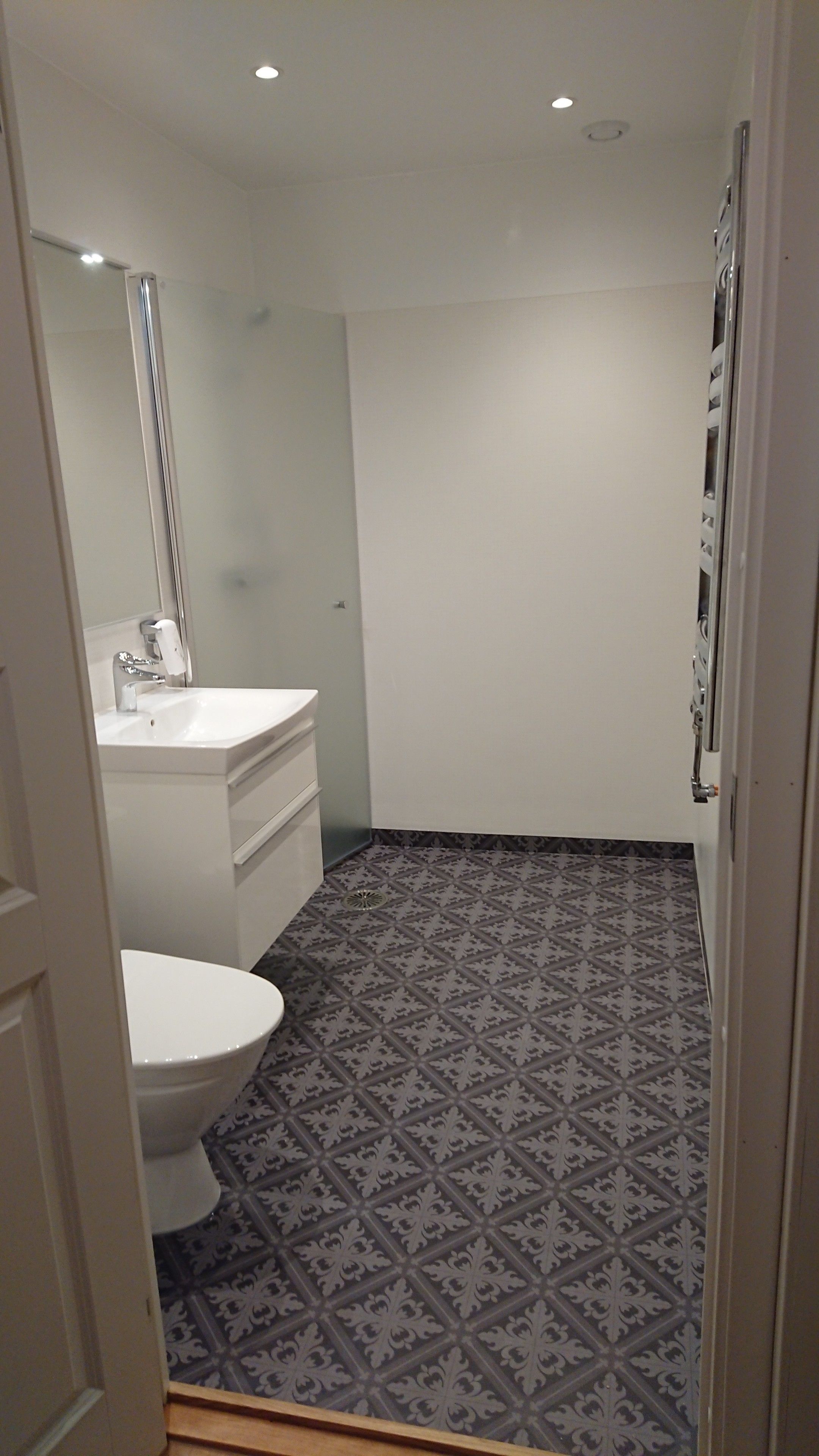 A small, modern bathroom with a white sink, mirror, and toilet. The floor has a patterned tile design.