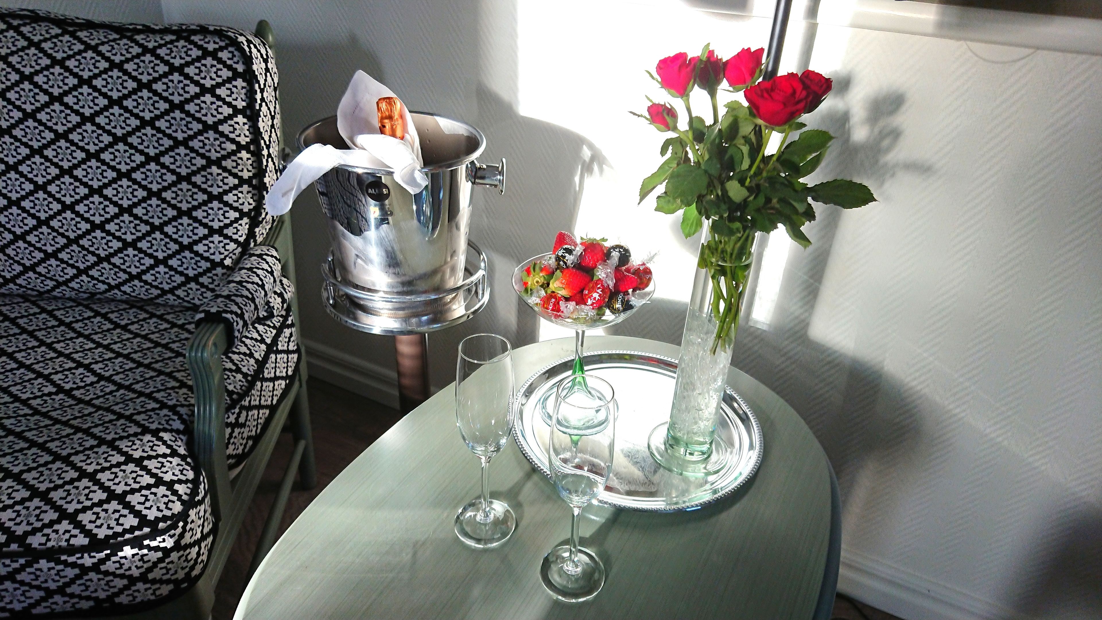 A table with a vase of red roses, a glass of water, and a tray with strawberries and chocolates.