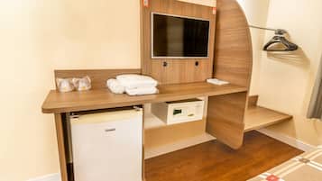 A compact hotel room with a wooden desk, a television, a mini-fridge, and a coat rack.