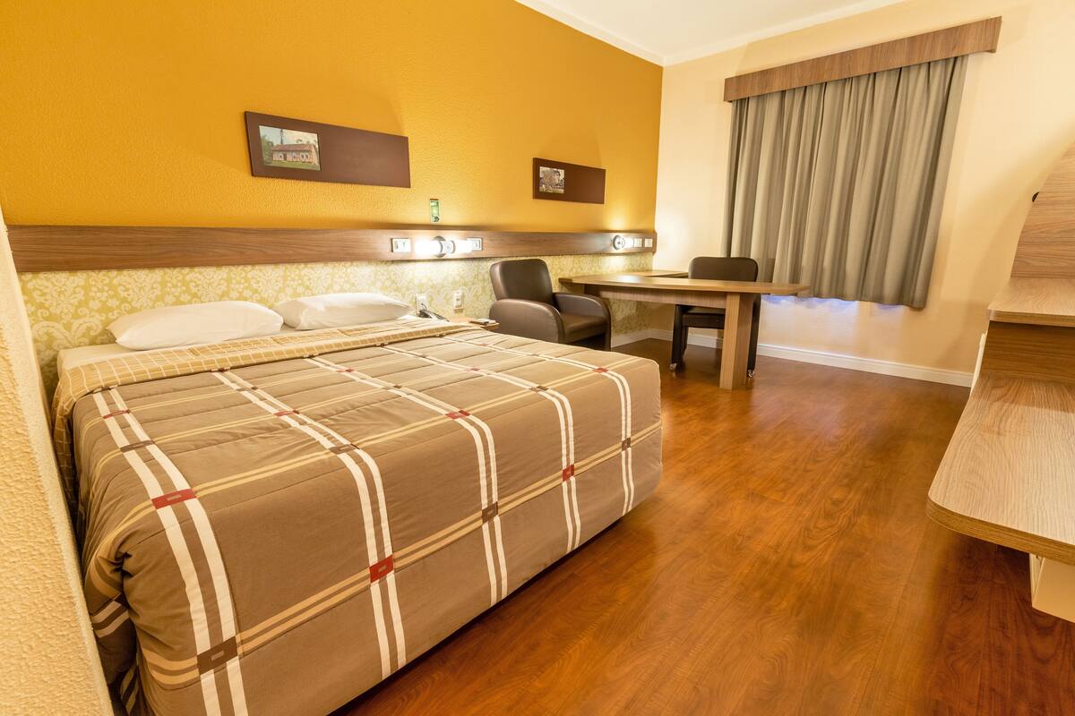 Una habitación de hotel con cama, escritorio, silla y ventana con cortinas.