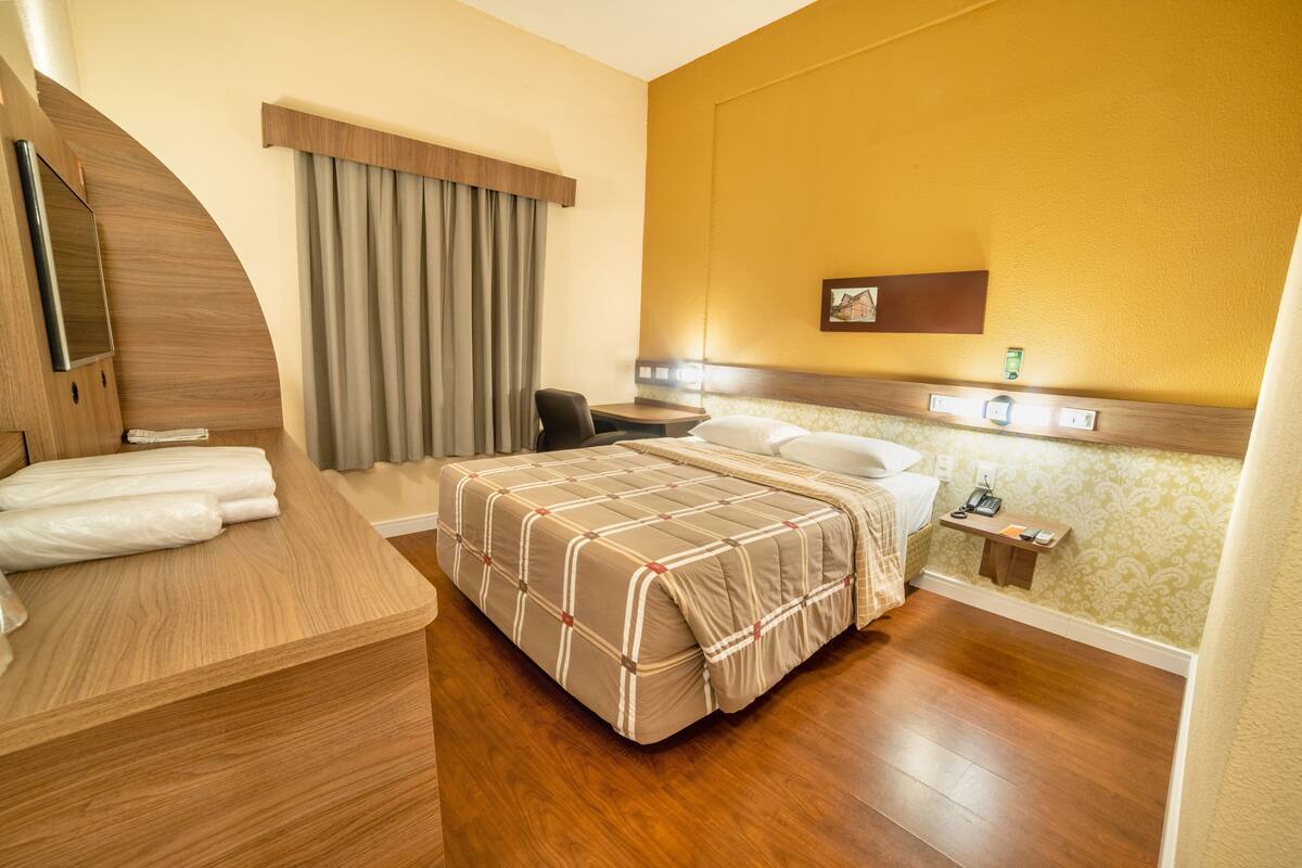 Habitación de hotel con cama, escritorio, silla, mesita, mesita de noche, espejo y ventana con cortinas.