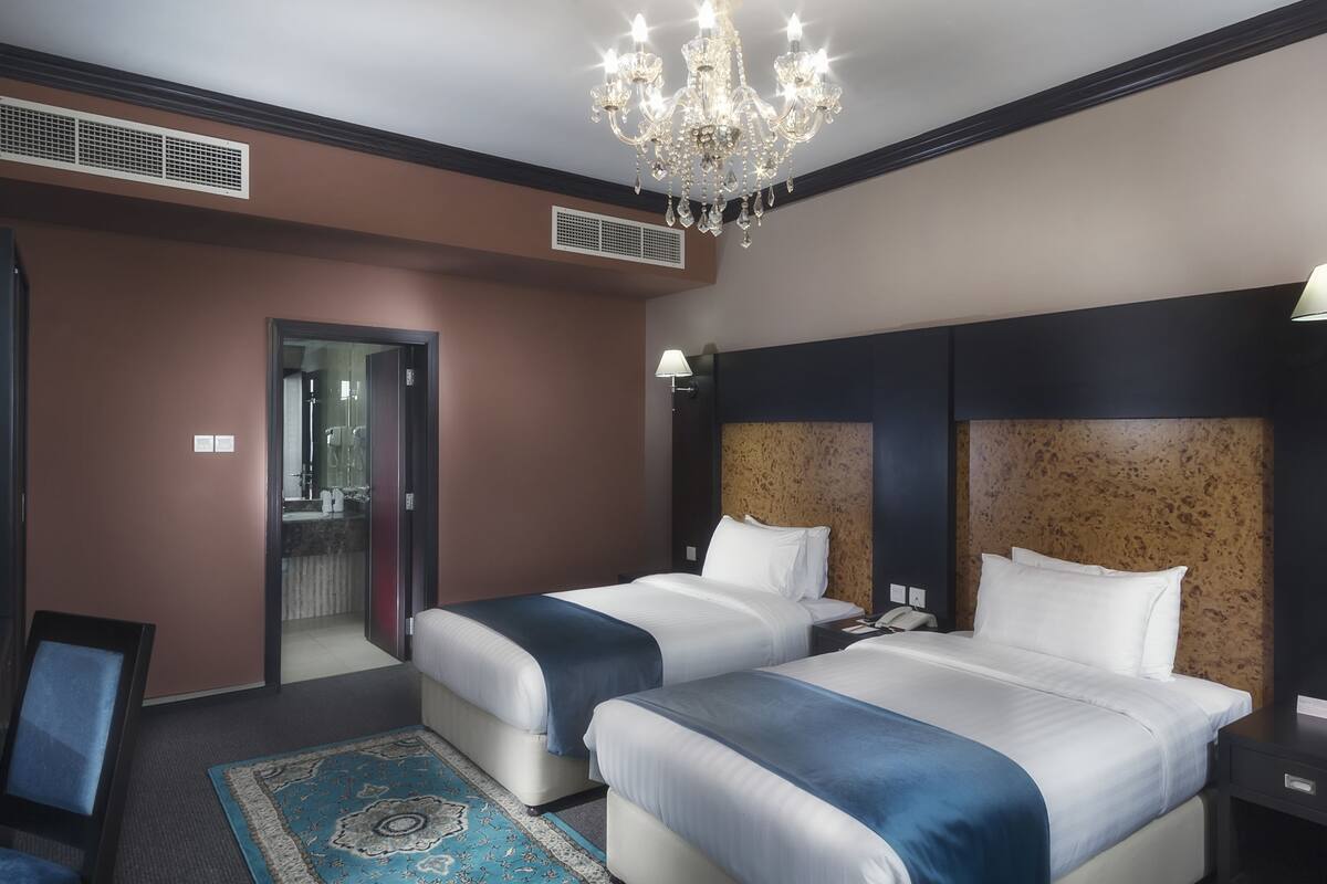 A hotel room with two beds, a chandelier, a bathroom visible through an open door, and a patterned rug.