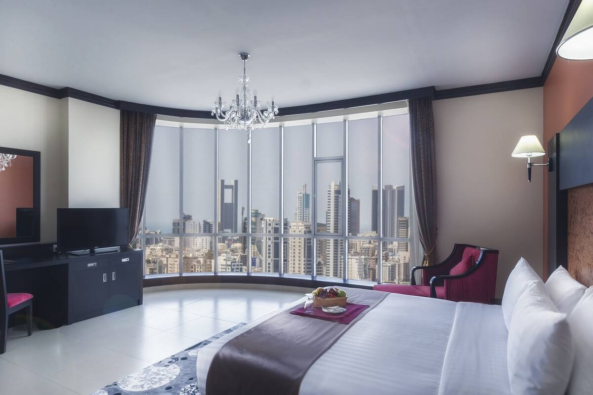 A hotel room with a large bed, a desk with a TV, a red chair, and a view of the cityscape through large windows.