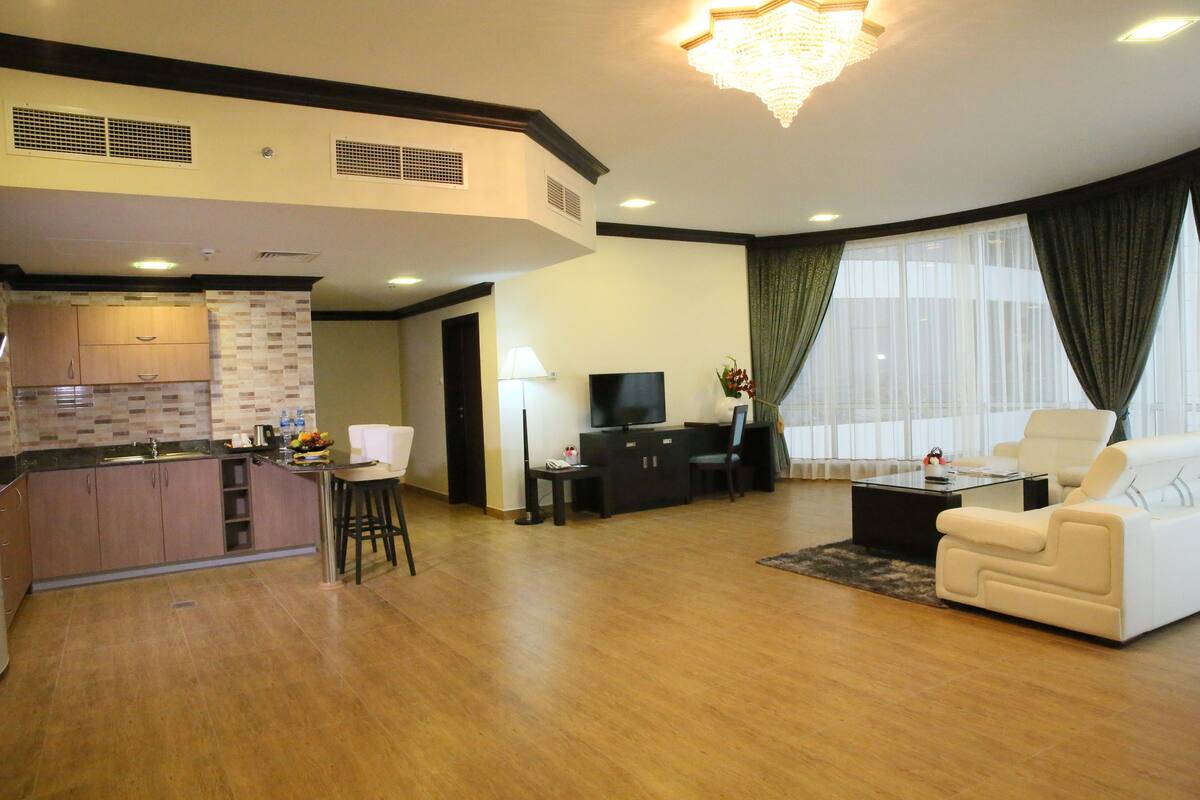 A modern living room with a sofa, a coffee table, and a TV. There is a kitchen area with a counter and cabinets.