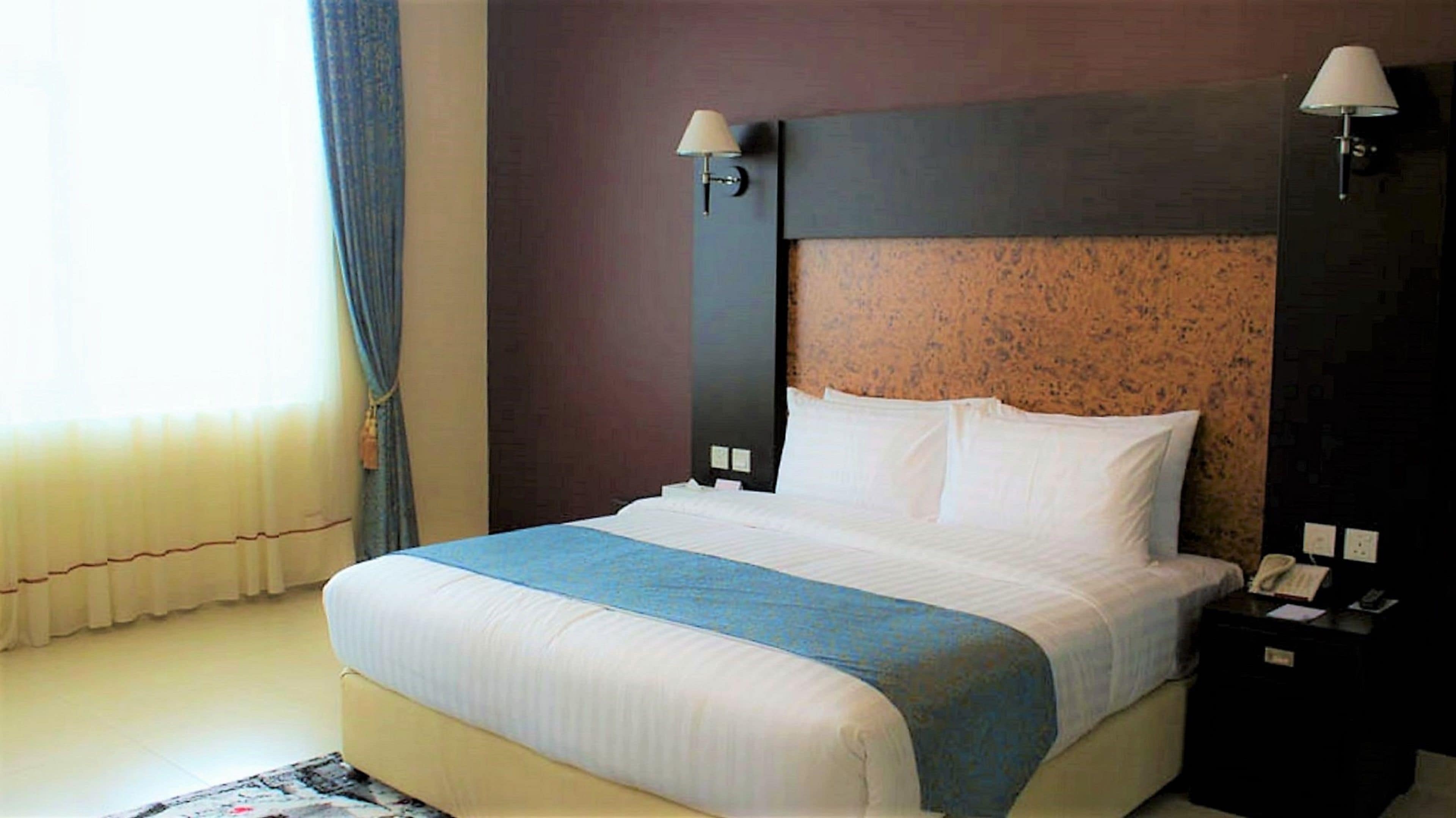 A hotel room with a large bed, two bedside lamps, a nightstand, and a window with curtains.