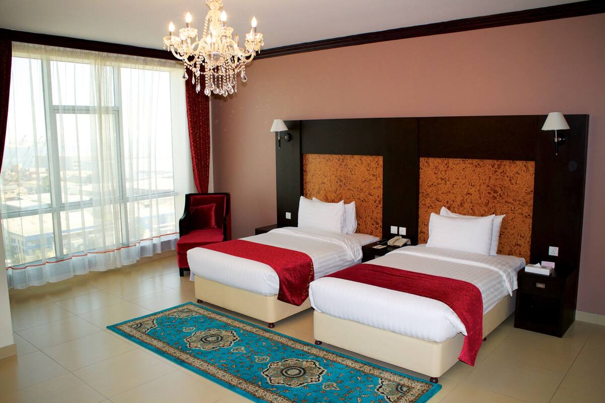 A hotel room with two beds, a chandelier, a large window with curtains, and a patterned rug.