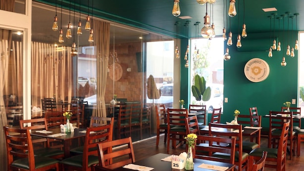 A restaurant interior with wooden tables and chairs, hanging light fixtures, and a green wall with a sign. - H+ Hotel (Dourados)