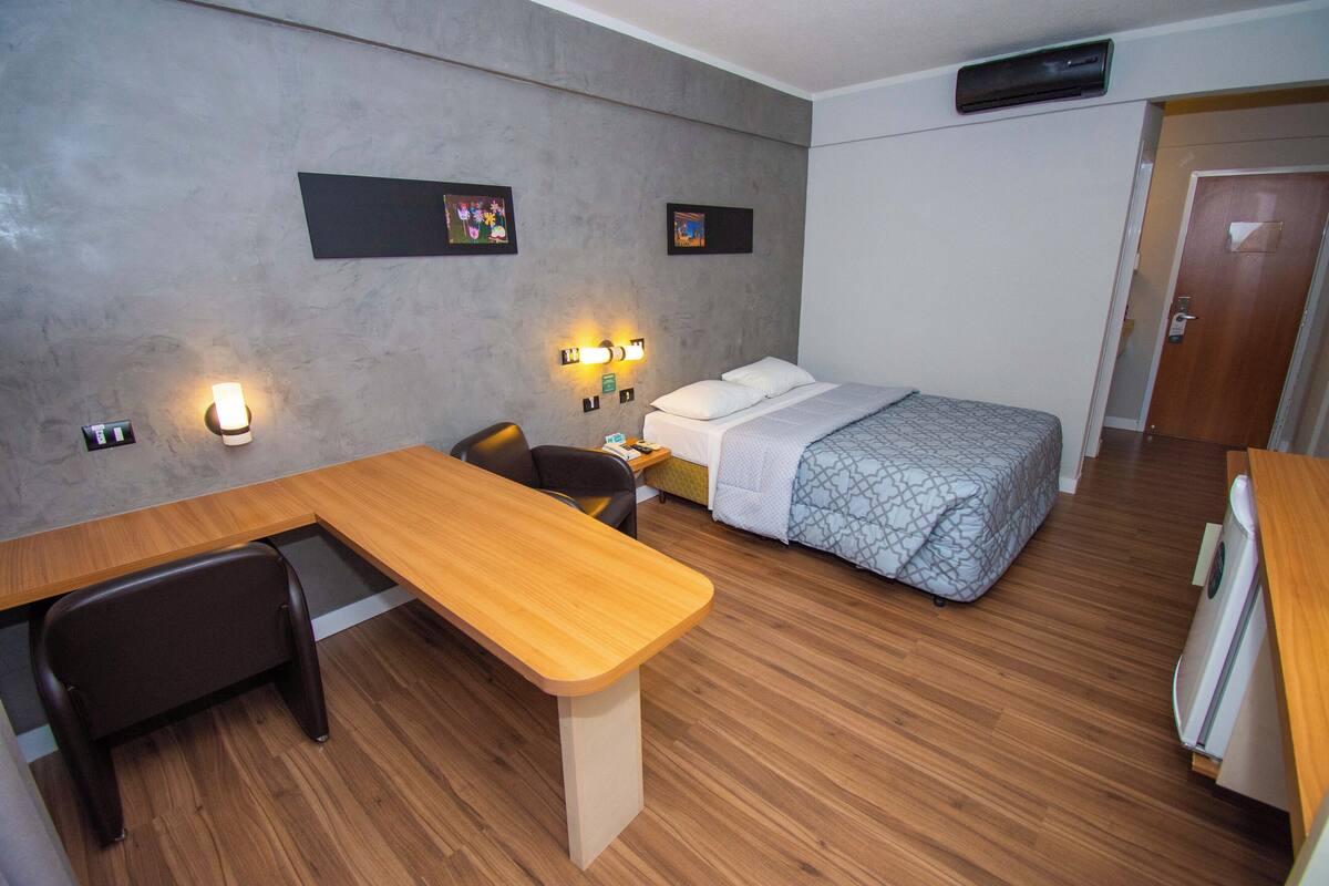A hotel room with a wooden desk, a bed with patterned bedding, a chair, and two wall-mounted light fixtures.