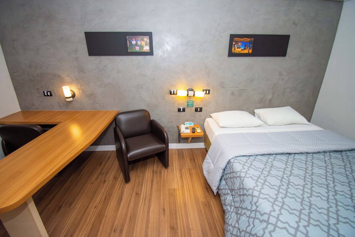 A hotel room with a wooden desk, a single chair, a bed with a patterned bedspread, and two wall-mounted light fixtures.