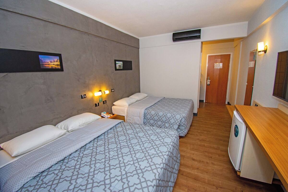 A hotel room with two beds, a wooden desk, a refrigerator, and a hallway leading to another room.