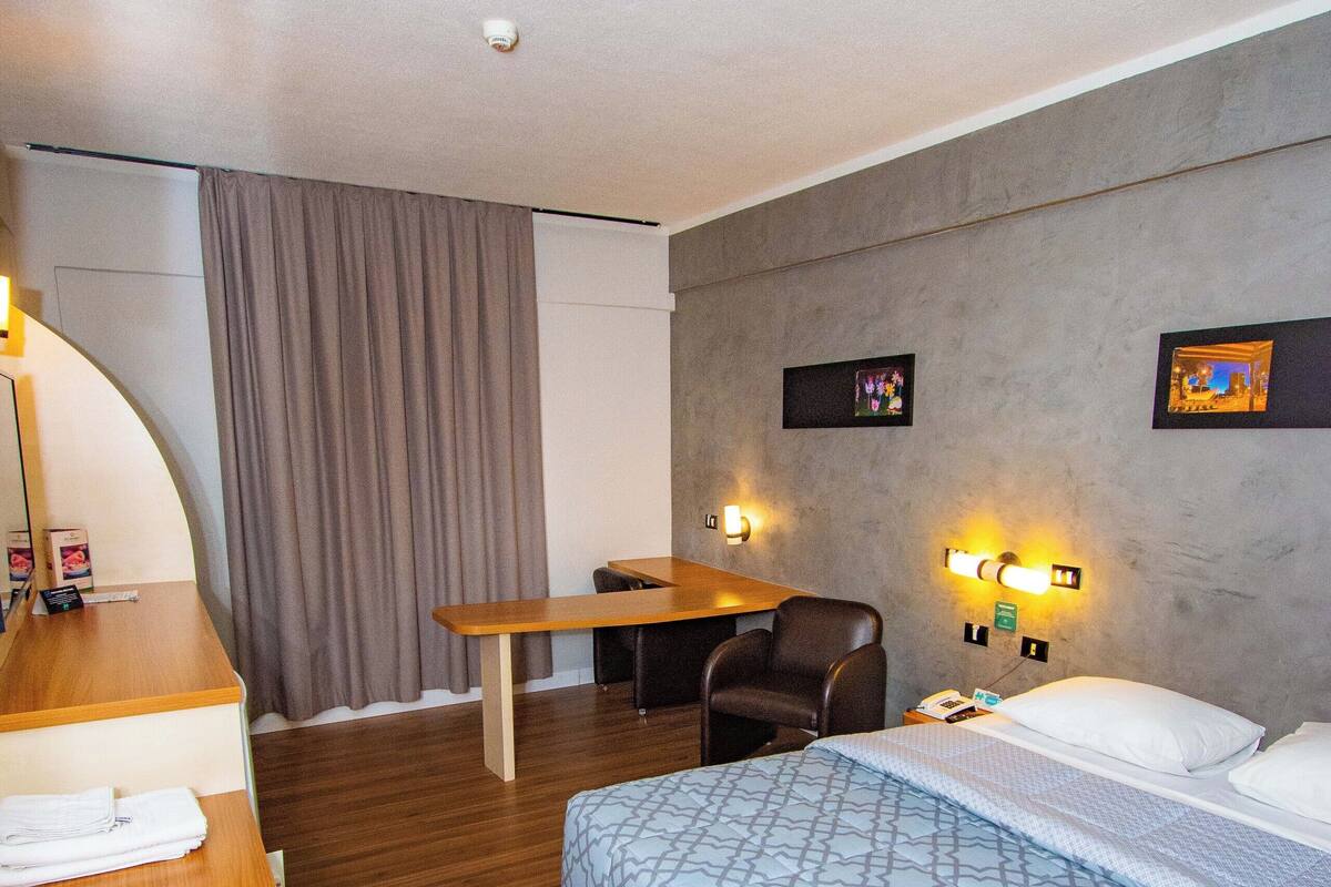 A hotel room with a bed, a desk, a chair, a table, and a wall-mounted light fixture.