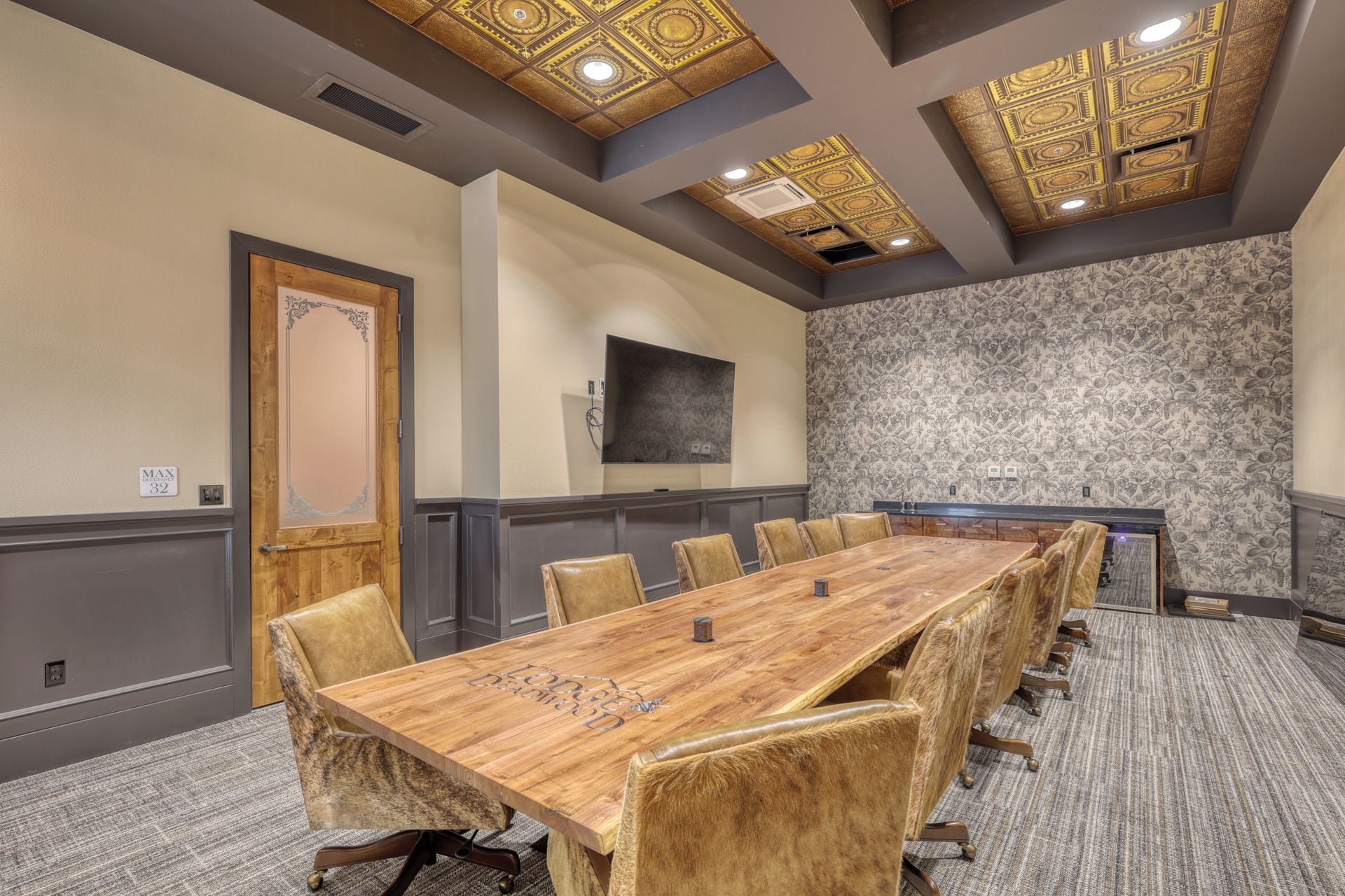 Un salón de conferencias con una larga mesa de madera, sillas de cuero, papel tapiz estampado y un techo decorativo.