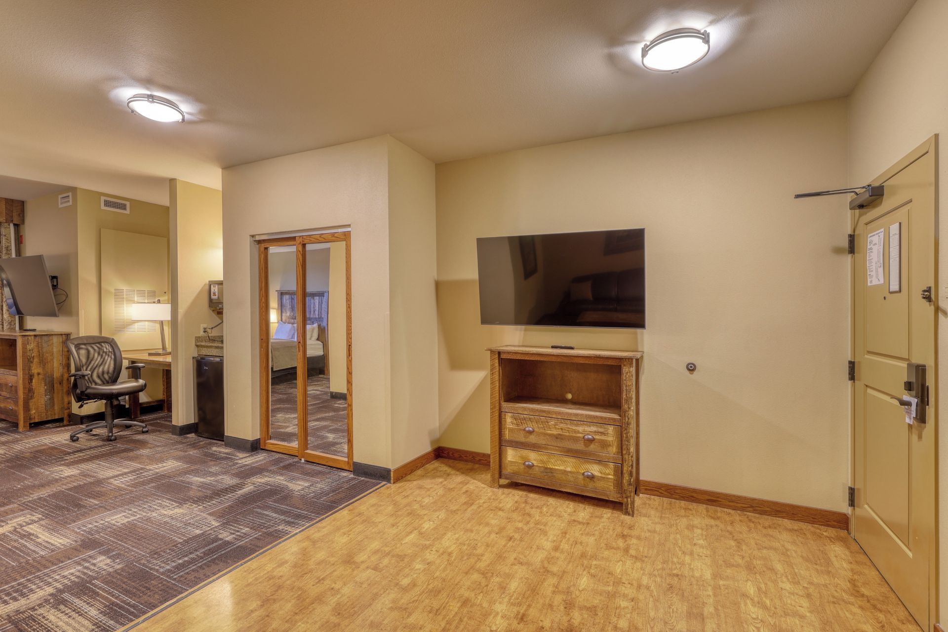 A hotel room with a flat-screen TV, a wooden dresser, a desk with a computer, and a carpeted floor.