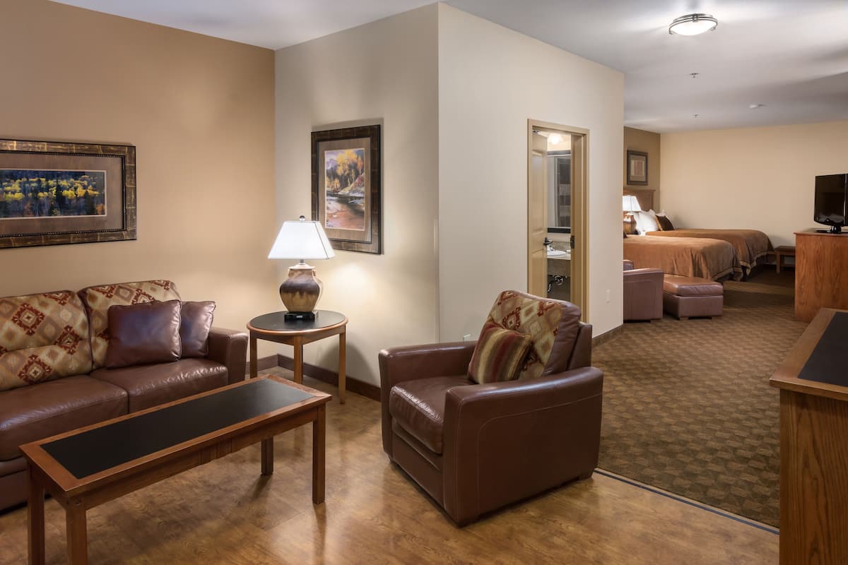 Habitación de hotel con zona de estar que incluye un sofá, un sillón, una mesita de centro y un cuadro en la pared. Se ve un baño a través de una puerta abierta y una cama.