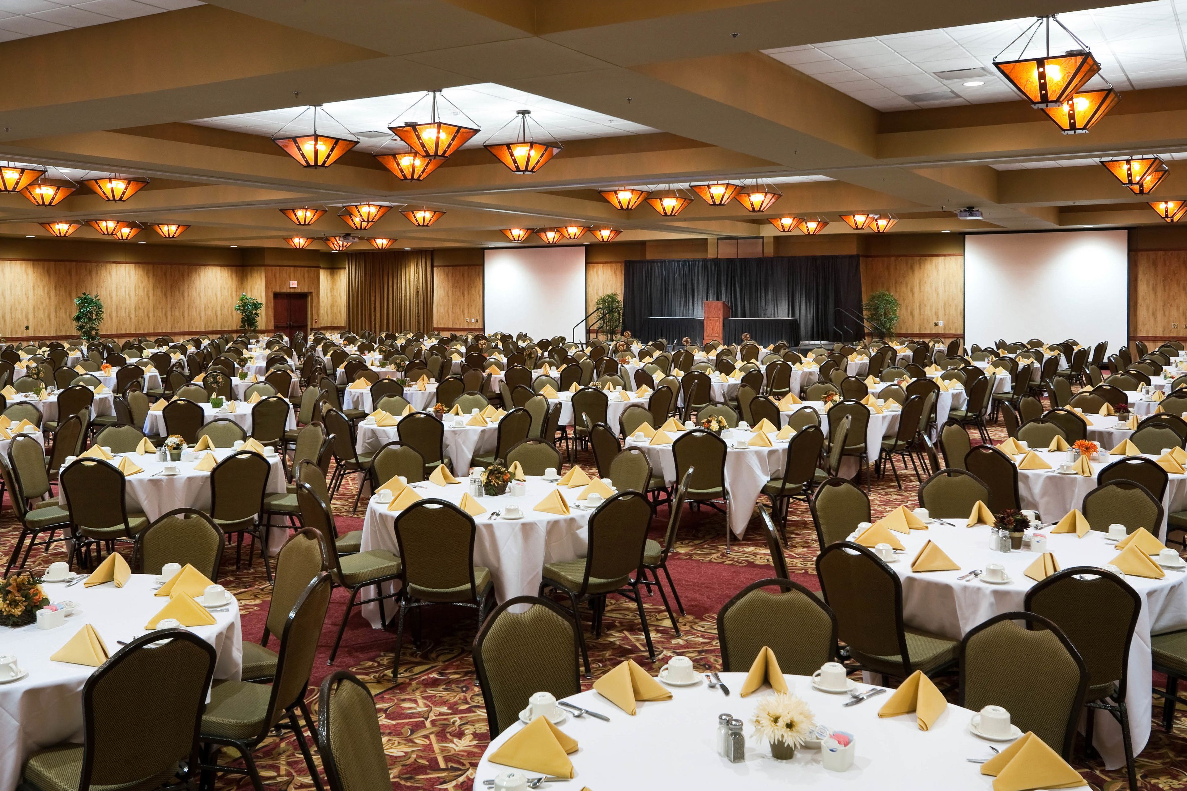 A large banquet hall with rows of tables set for a formal event, equipped with chandeliers and a stage.