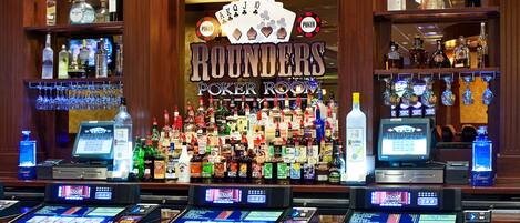 A poker room bar with multiple gaming tables, a variety of liquor bottles, and a neon sign.