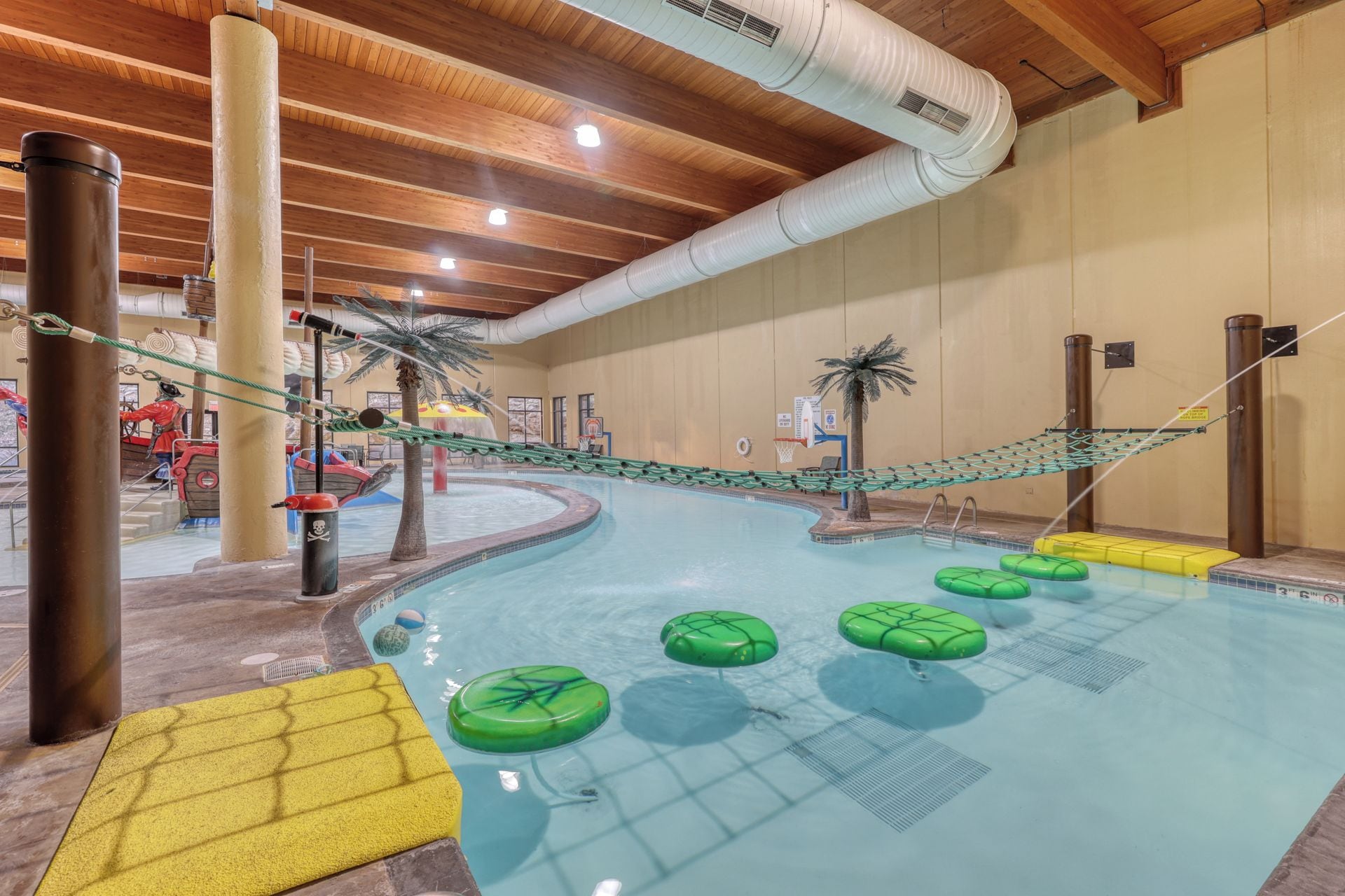 An indoor swimming pool with a rope bridge, palm trees, and green floating platforms.