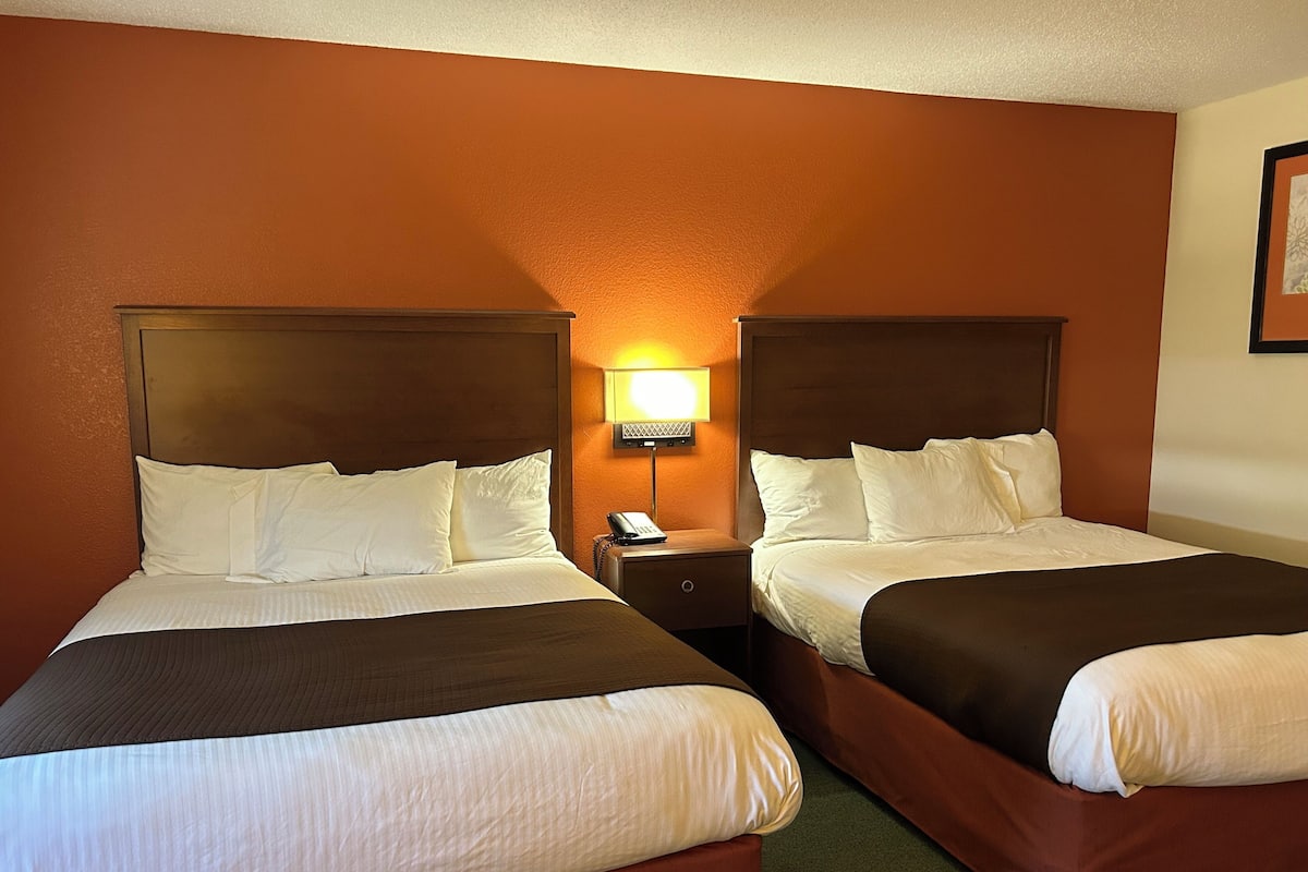 Quarto de hotel com duas camas, cada uma com cabeceira, mesa de cabeceira com abajur e um quadro na parede.