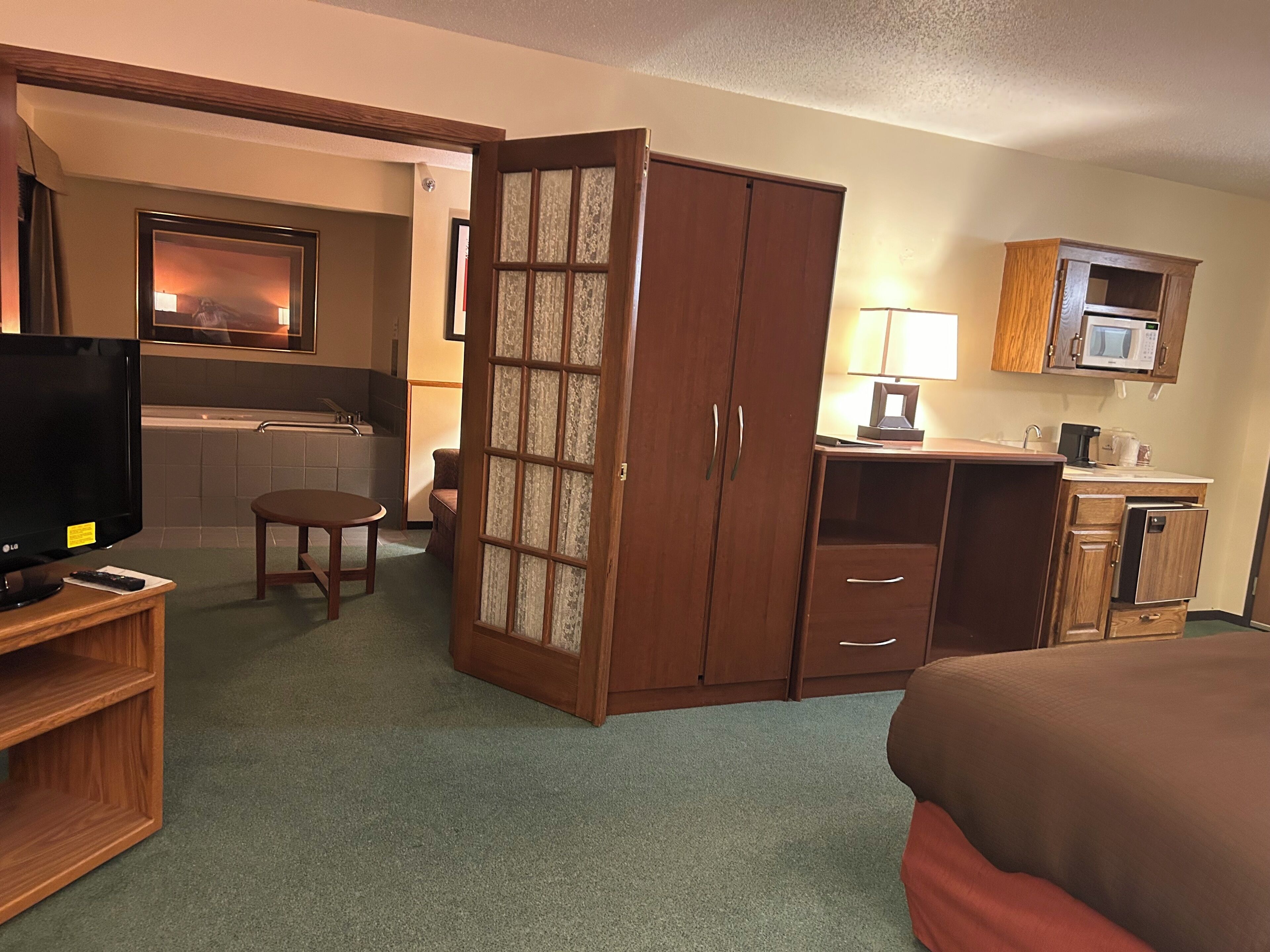 Habitación de hotel con cama, televisor, escritorio con lámpara, microondas y pequeña cocina.